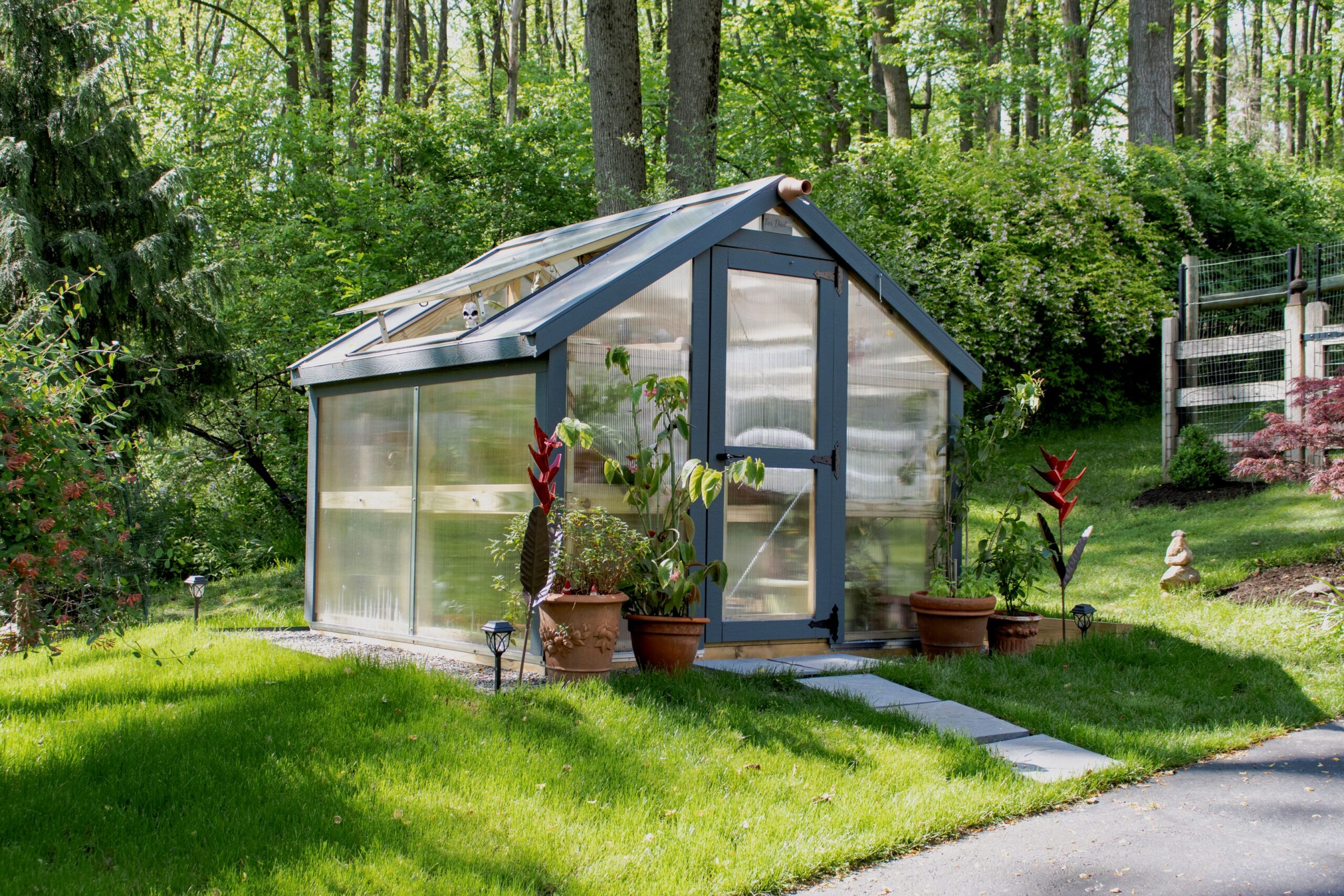 Left side view of a gray A-Frame Greenhouse with stone pathway and potted plants by the door