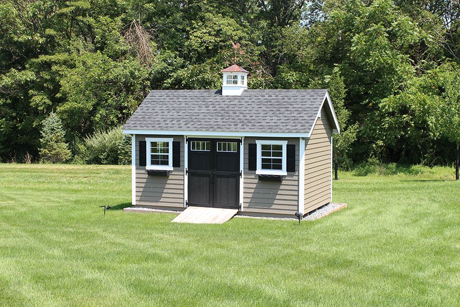 Garden shed with tan vinyl siding, white trim, black double doors with windows, gray roofing with a copper cupola, and white windows with black shutters and black window boxes.
