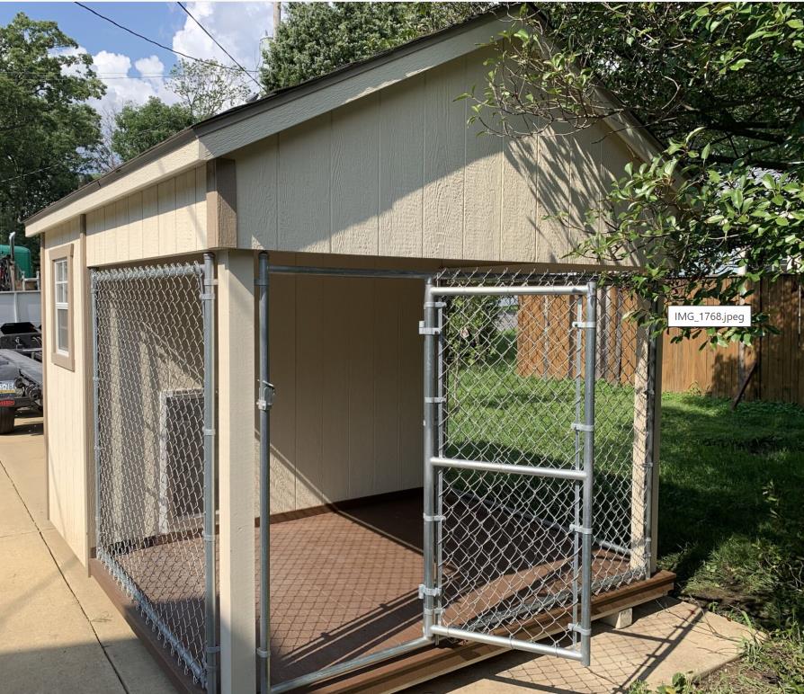 Front exterior view of an 8x10 residential traditional dog kennel with light tan wood siding, brown trim, and a chain link enclosed dog run with an open door.