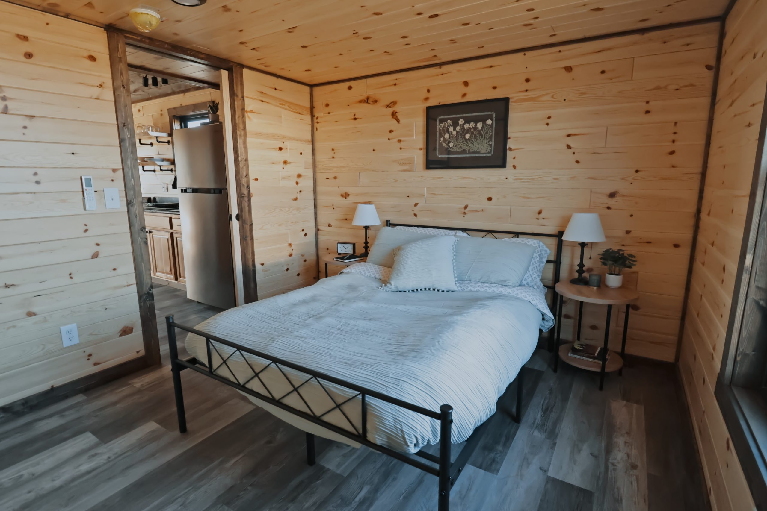 Bedroom of an 1889 Park Model with natural wood walls, gray vinyl plank flooring, brown stained trim, a black iron bed with white bedding, and round bedside tables with lamps.