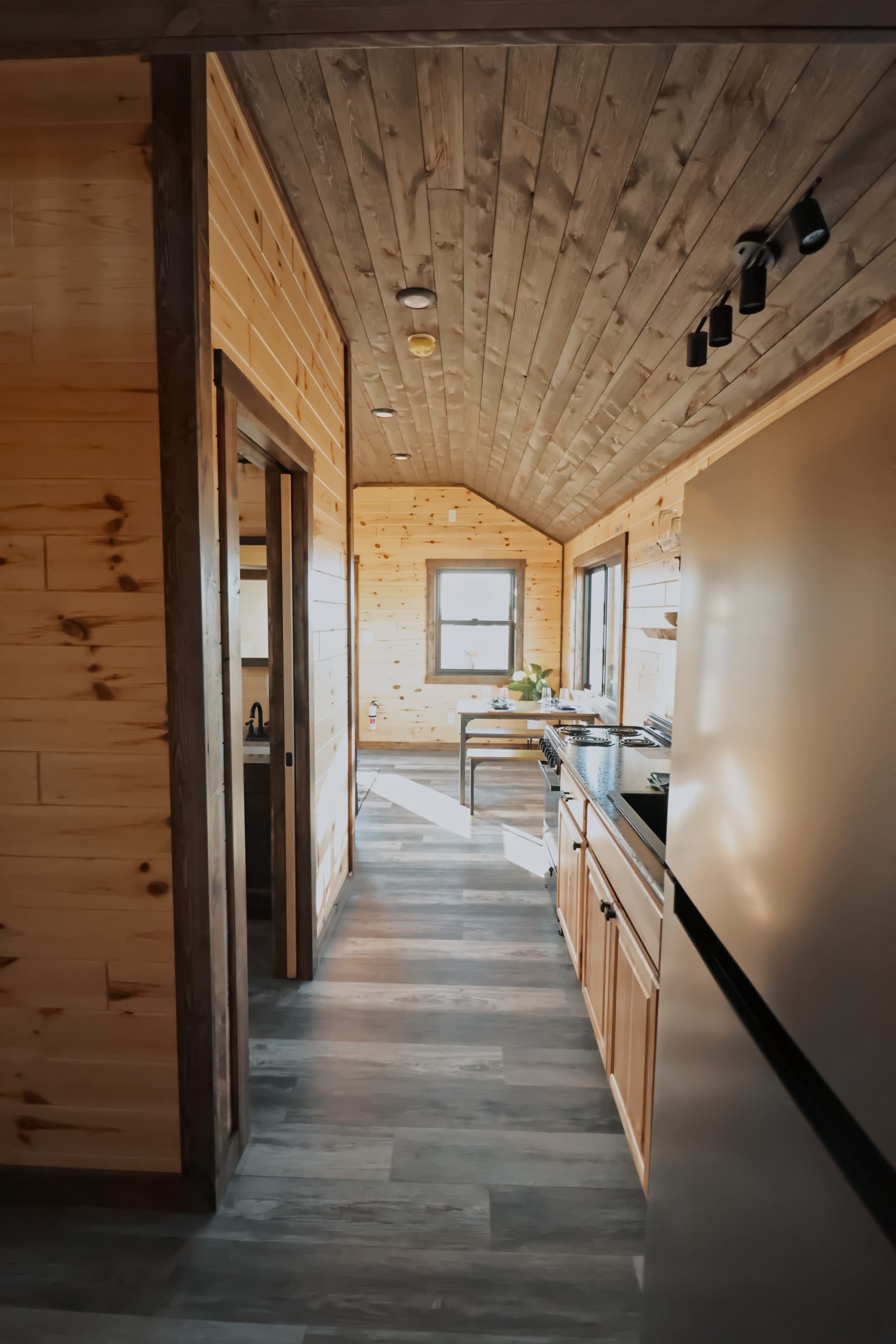 Hallway leading into a kitchen in an 1889 Park Model with natural wood walls, brown stained trim, gray vinyl plank flooring, and a brown plank ceiling.