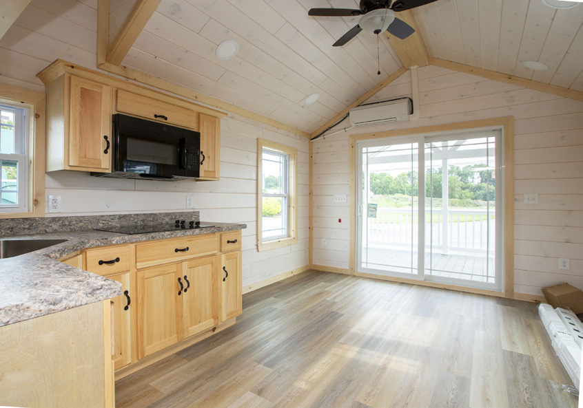 Interior view of an Appalachian Park Model living room and kitchen with white wood panel walls, vinyl plank flooring, a wall-mounted air conditioning unit, light colored wood kitchen cabinets, a black microwave, and a ceiling fan.