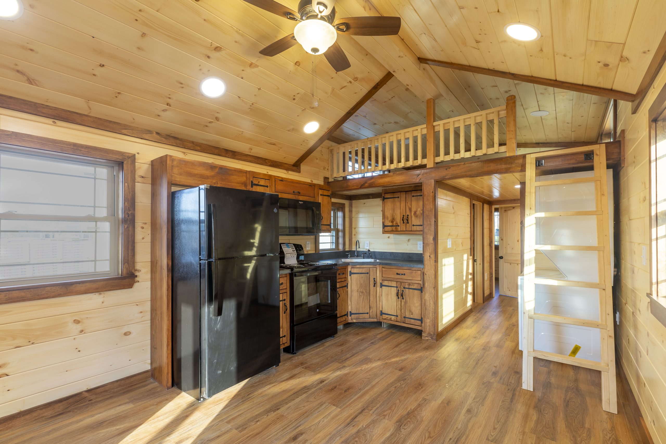 Interior view of an Appalachian Park Model with natural wood walls, a loft and ladder, stained wood kitchen cabinets, black kitchen appliances, and vinyl plank flooring.