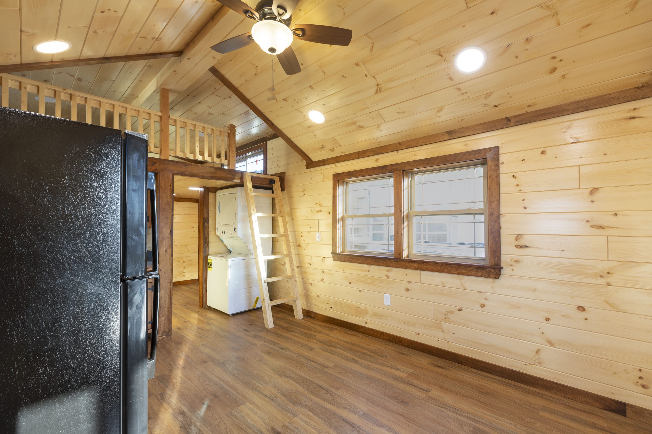 Interior view of an Appalachian Park Model with natural wood walls, a loft and ladder, a double window with stained trim, a black fridge, a stacked washer and dryer, and vinyl plank flooring.