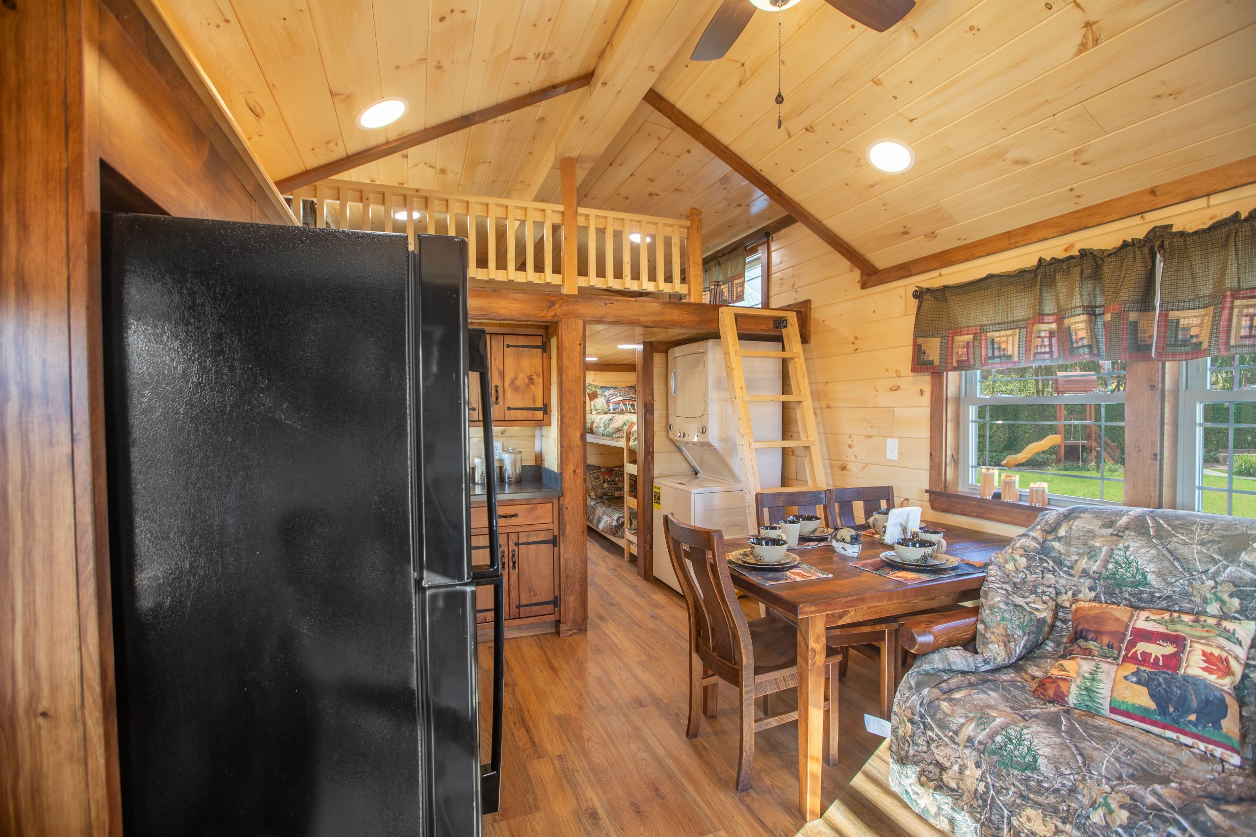 Finished interior of an Appalachian Park Model with natural wood walls, a loft and ladder, a double window with green half curtains, a black fridge, a stacked washer and dryer, a wooden dining table and chairs, a camo couch, and vinyl plank flooring.