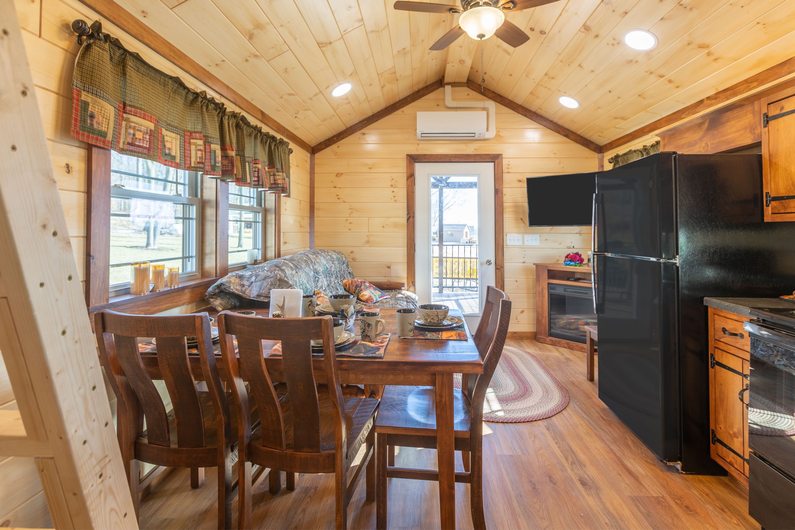 Finished interior of an Appalachian Park Model with natural wood walls, a single glass door entrance, a double window with green half curtains, a black fridge, a stacked washer and dryer, a wooden dining table and chairs, a camo couch, a corner tv over an electric fireplace, and vinyl plank flooring.