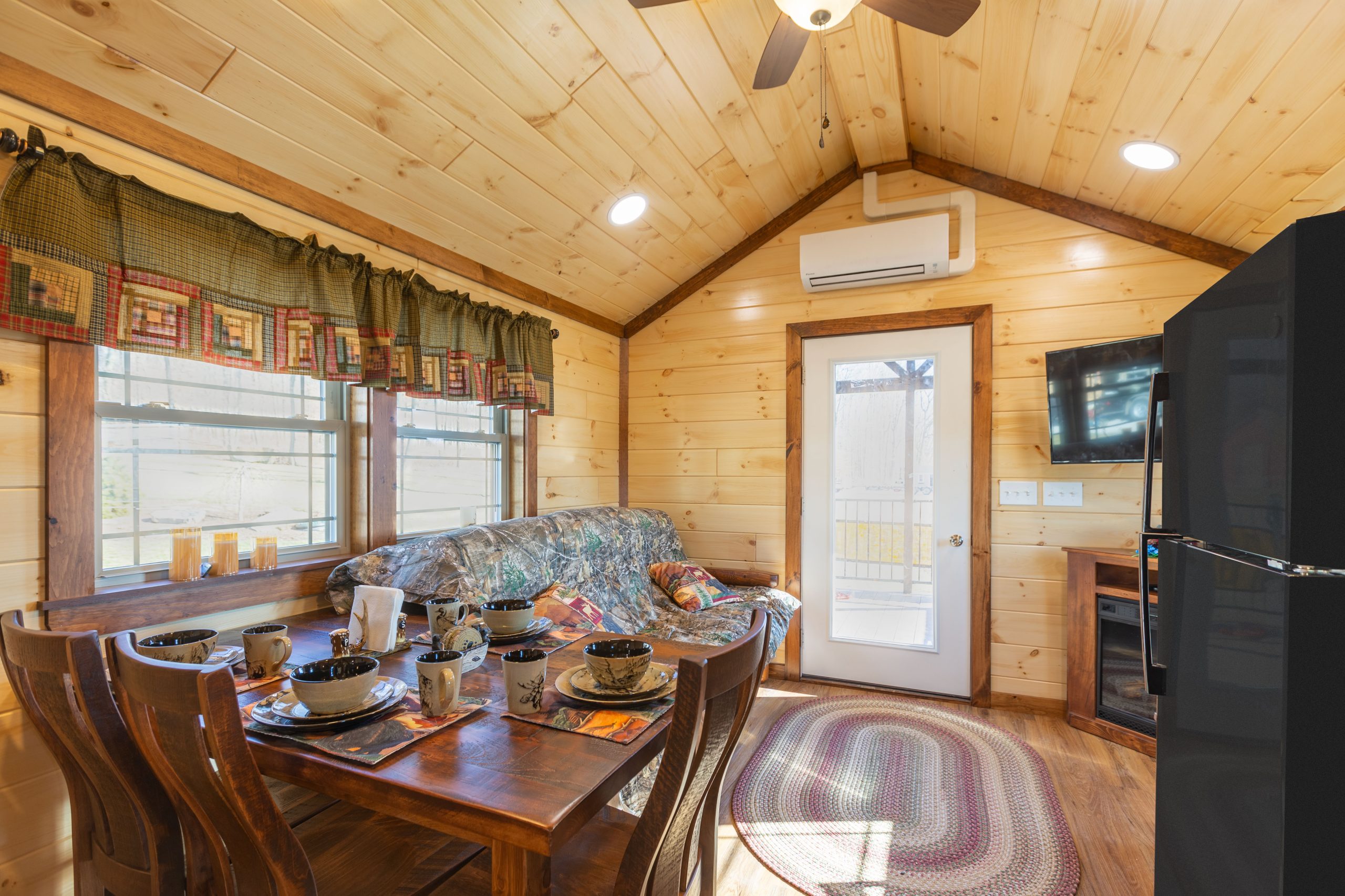 Finished interior of an Appalachian Park Model with natural wood walls, a single glass door entrance, a double window with green half curtains, a black fridge, a stacked washer and dryer, a wooden dining table and chairs, a camo couch, a corner tv over an electric fireplace, and vinyl plank flooring.