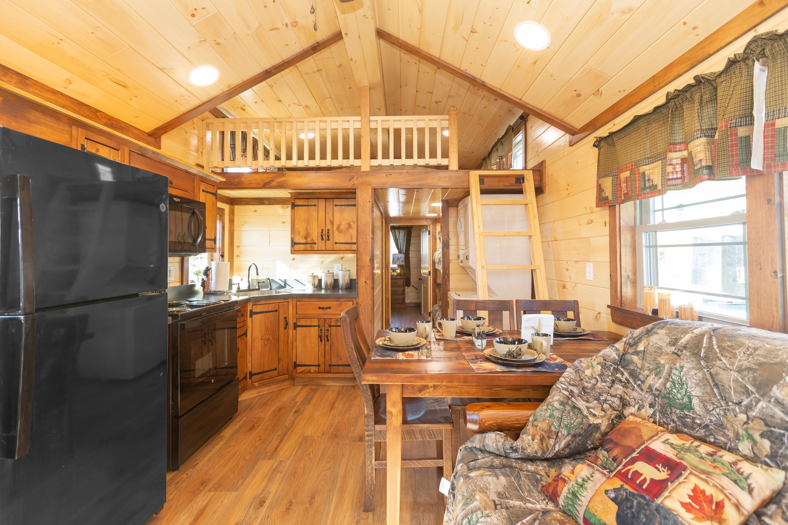 Finished interior of an Appalachian Park Model with natural wood walls, a loft and ladder, a double window with green half curtains, stained wood kitchen cabinets, black kitchen appliances, a stacked washer and dryer, a wooden dining table and chairs, a camo couch, and vinyl plank flooring.