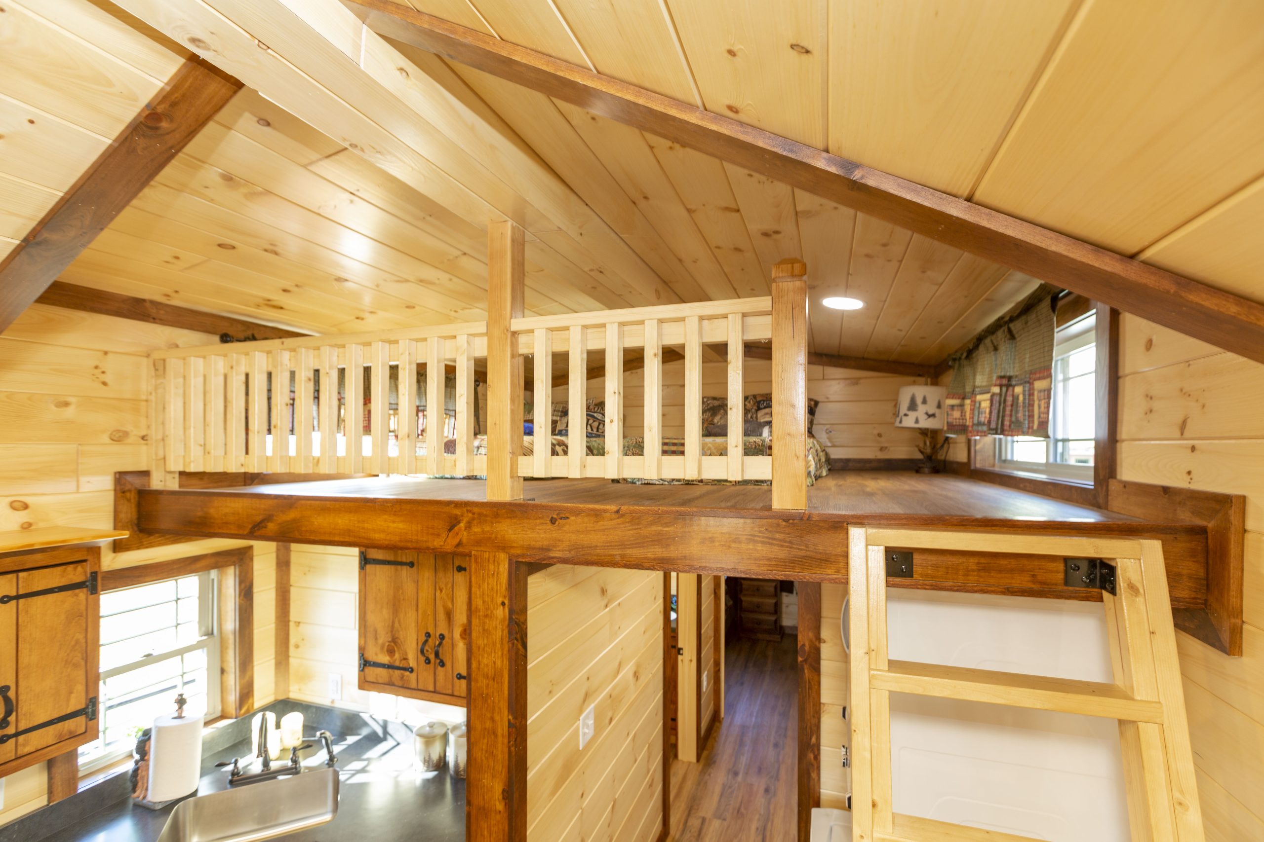 Close up of an Appalachian Park Model loft with natural wood walls, windows with half curtains, a lamp, and 2 single beds.