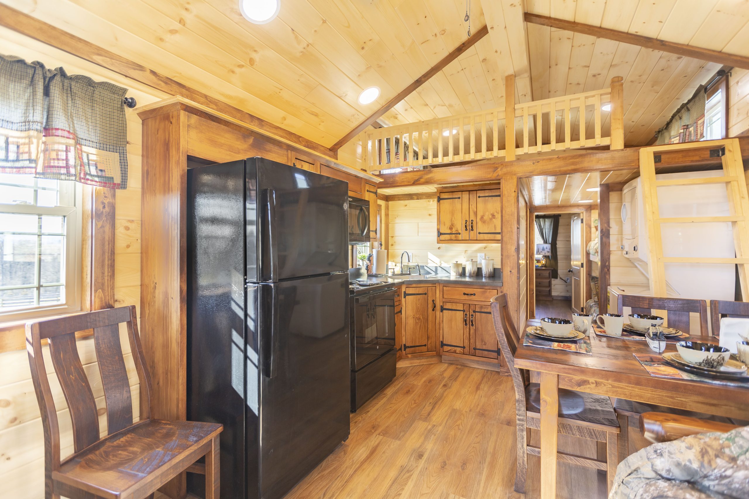 Finished interior of an Appalachian Park Model with natural wood walls, a loft and ladder, a double window with green half curtains, stained wood kitchen cabinets, black kitchen appliances, a stacked washer and dryer, a wooden dining table and chairs, and vinyl plank flooring.