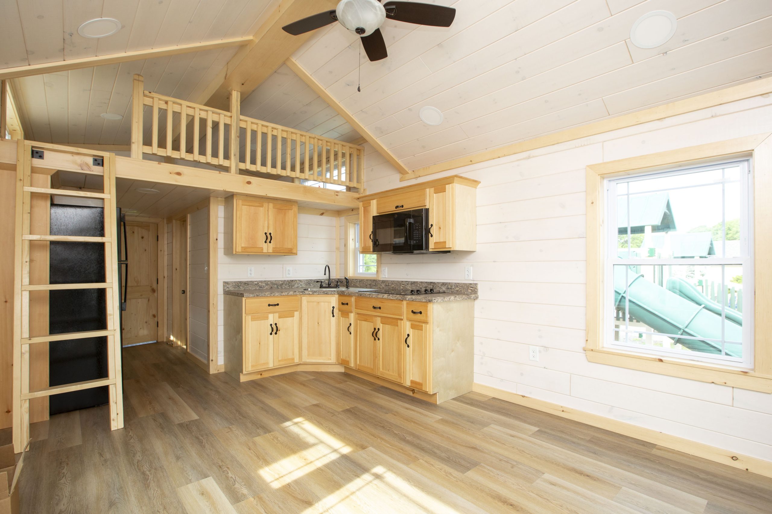 Interior view of an Appalachian Park Model with a loft, ladder, white wood panel walls, vinyl plank floors, and light wood kitchen cabinets.