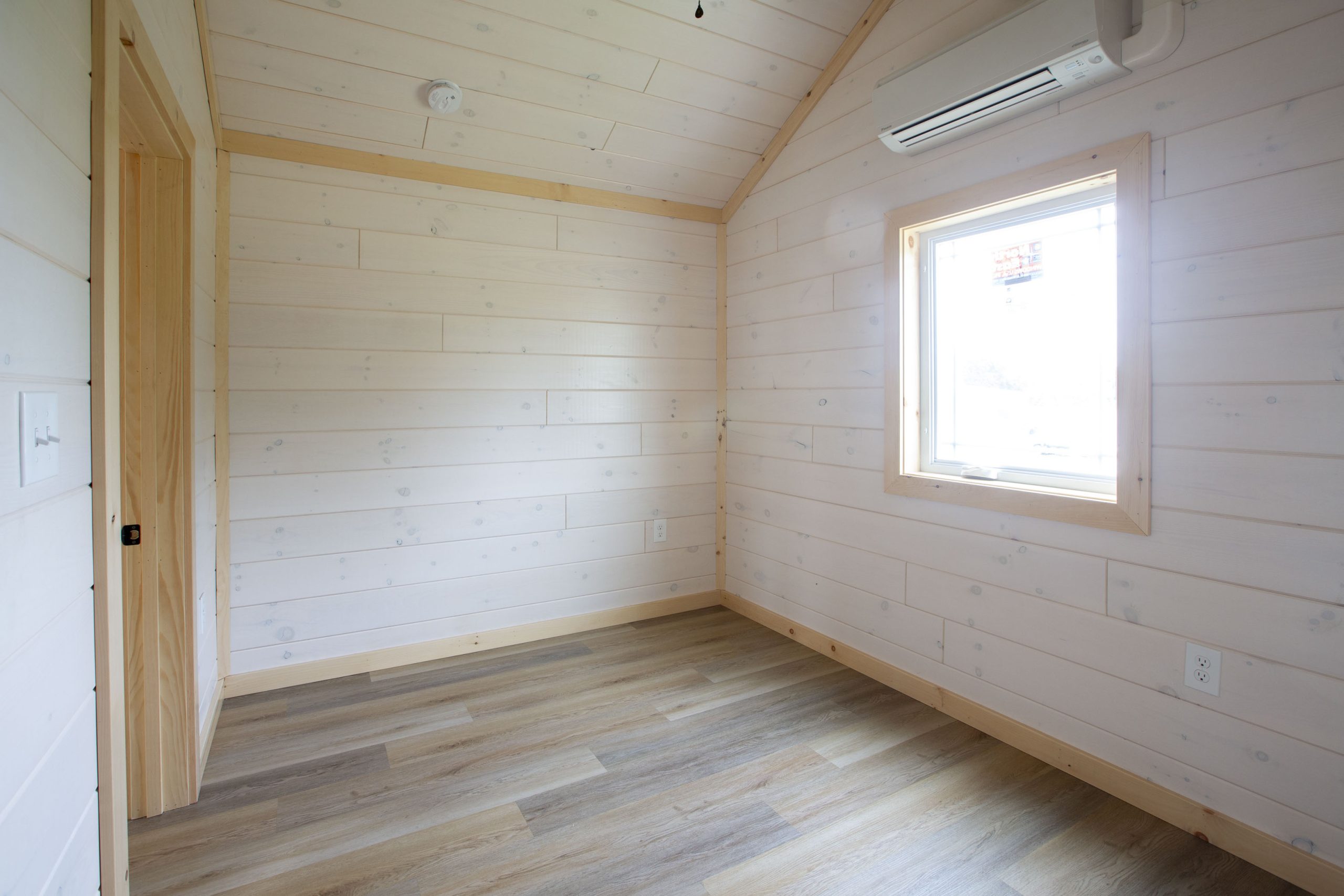 View of a room in an Appalachian Park Model with white wood wall panels, a wall mounted air conditioning unit, a square window, natural wood trim, and vinyl plank flooring.