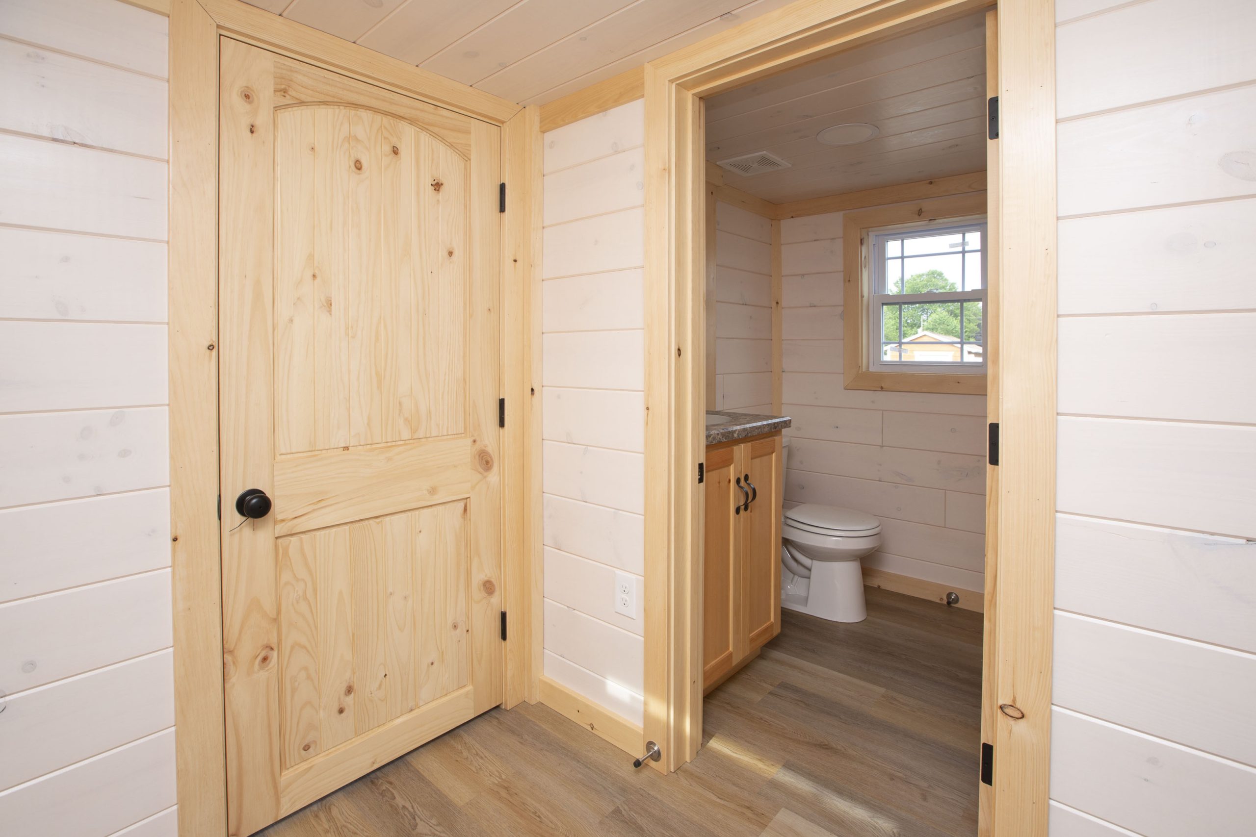 Hallway view of an Appalachian Park Model with white wood panel walls, a closed natural wood door, and an open door leading into a bathroom.