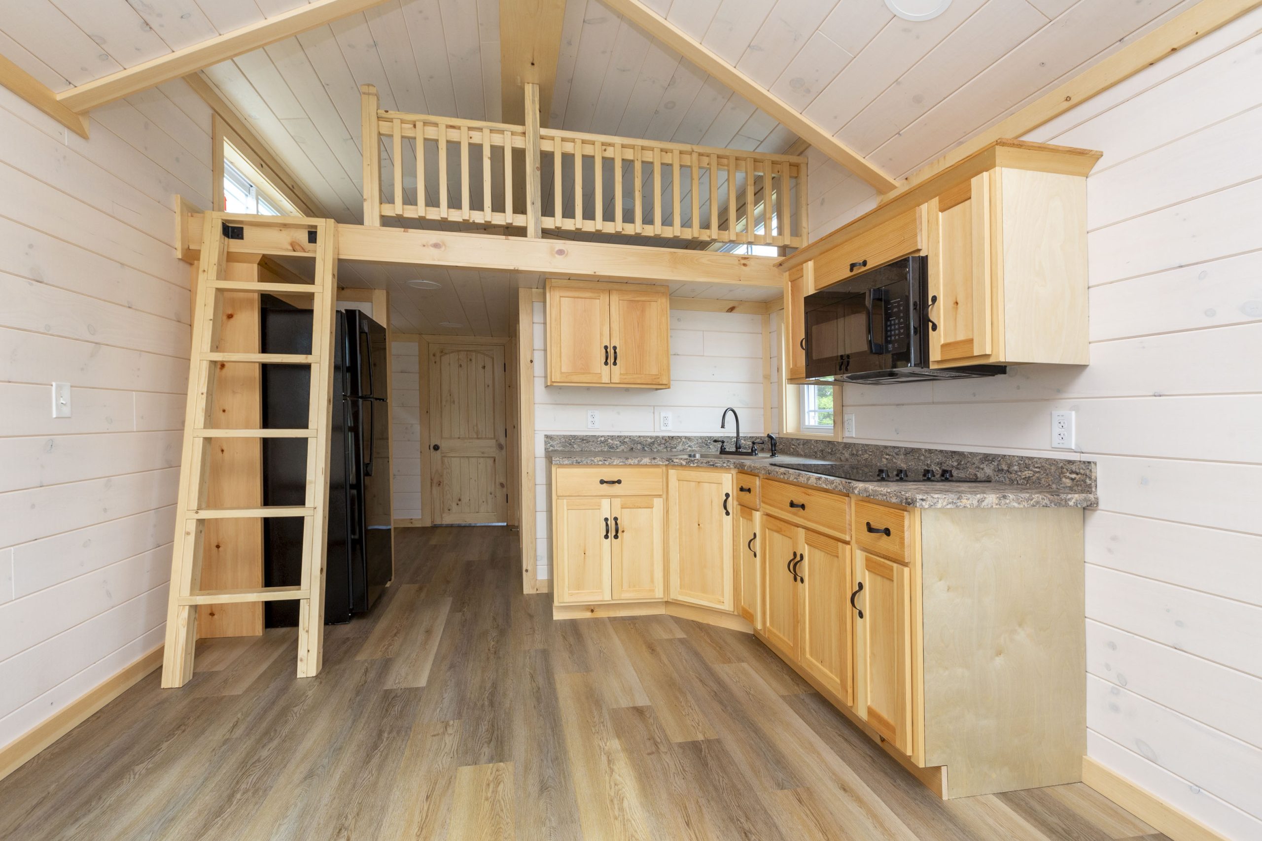 Overview of an Appalachian Park Model kitchen with light colored cabinets, a black microwave, white wood panel walls, vinyl plank flooring, and a loft with a ladder.