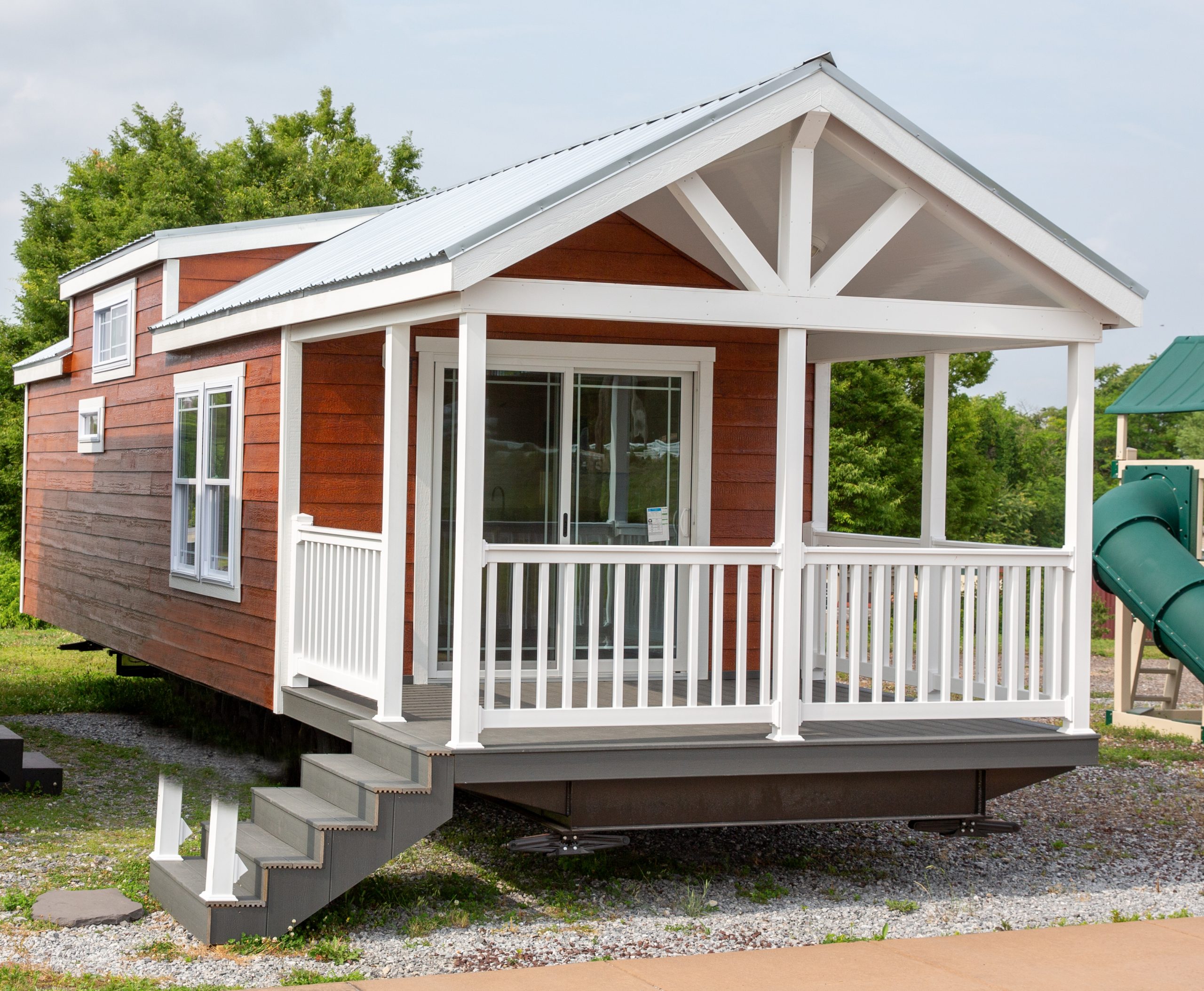 Front exterior view of an Appalachian Park Model with stained wood siding, white trim, white windows, gray steps, and a white covered front porch.