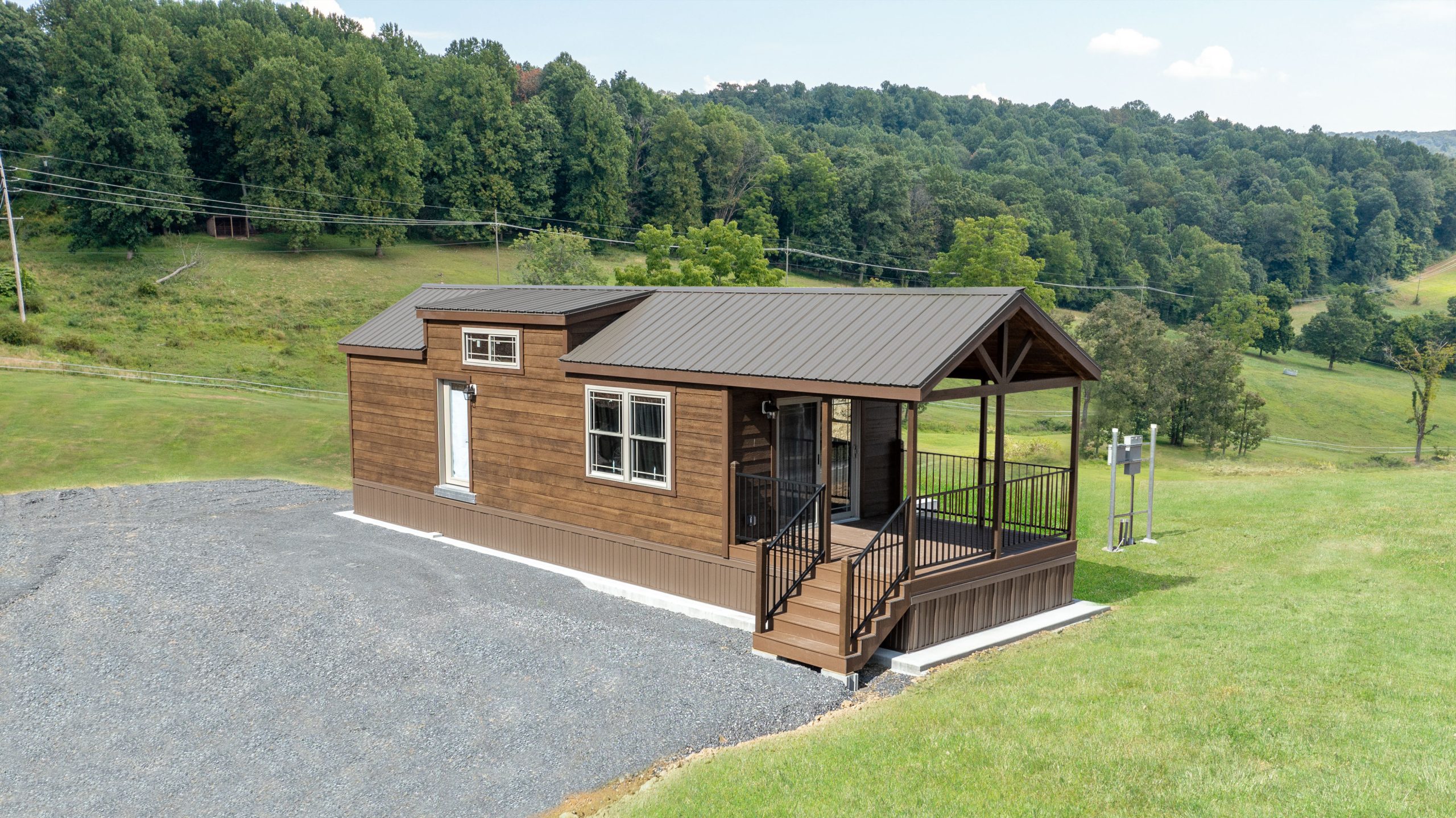 Front exterior of a Pine Cone Park Model with stained wood siding, brown trim, a brown metal roof, a fenced-in front porch, white windows, and white glass double doors.