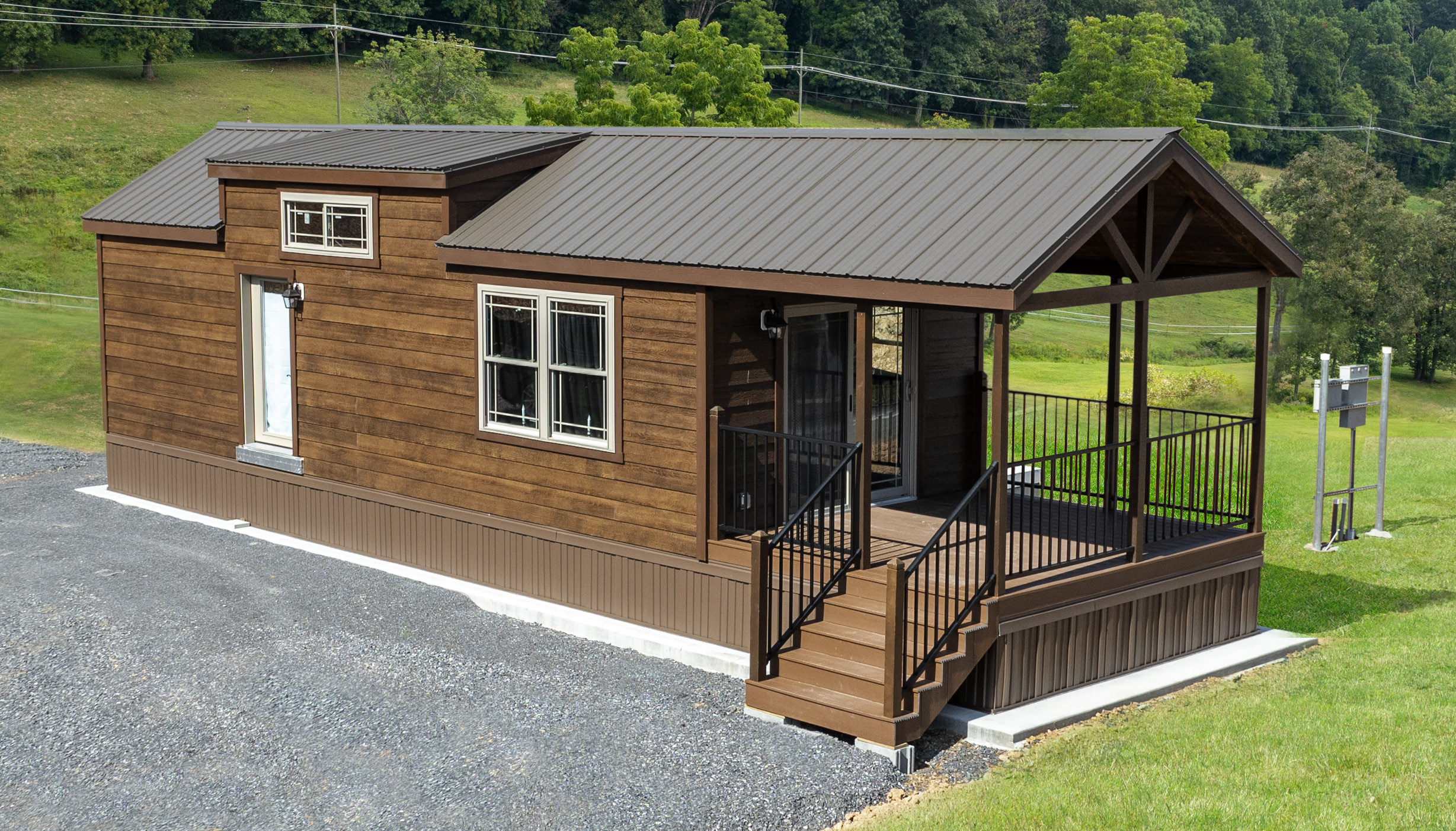 Front exterior of a Pine Cone Park Model with stained wood siding, brown trim, a brown metal roof, a fenced-in front porch, white windows, and white glass double doors.