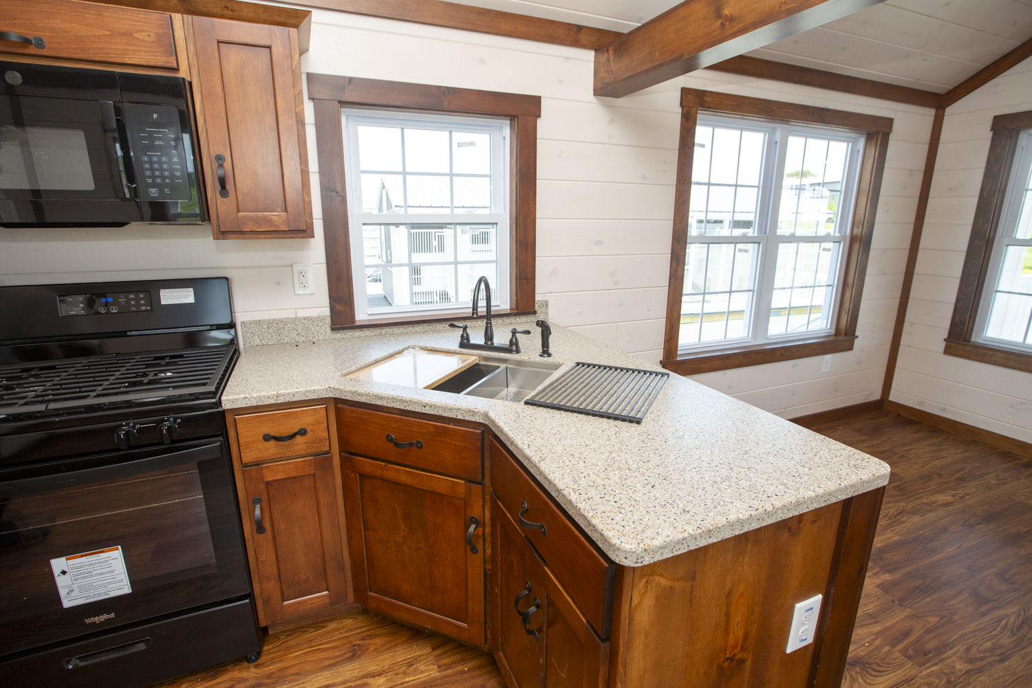 Part of a Santa Rosa Park Model kitchen with brown stained wood cabinets, black appliances, a large kitchen sink, white wood plank walls, and white windows with brown trim.