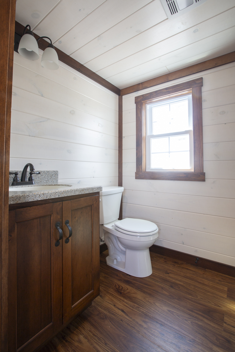 A bathroom in a Santa Rosa Park Model with white plank walls, dark vinyl plank flooring, a window, a white toilet, and a dark wood sink vanity with a dark metallic faucet.