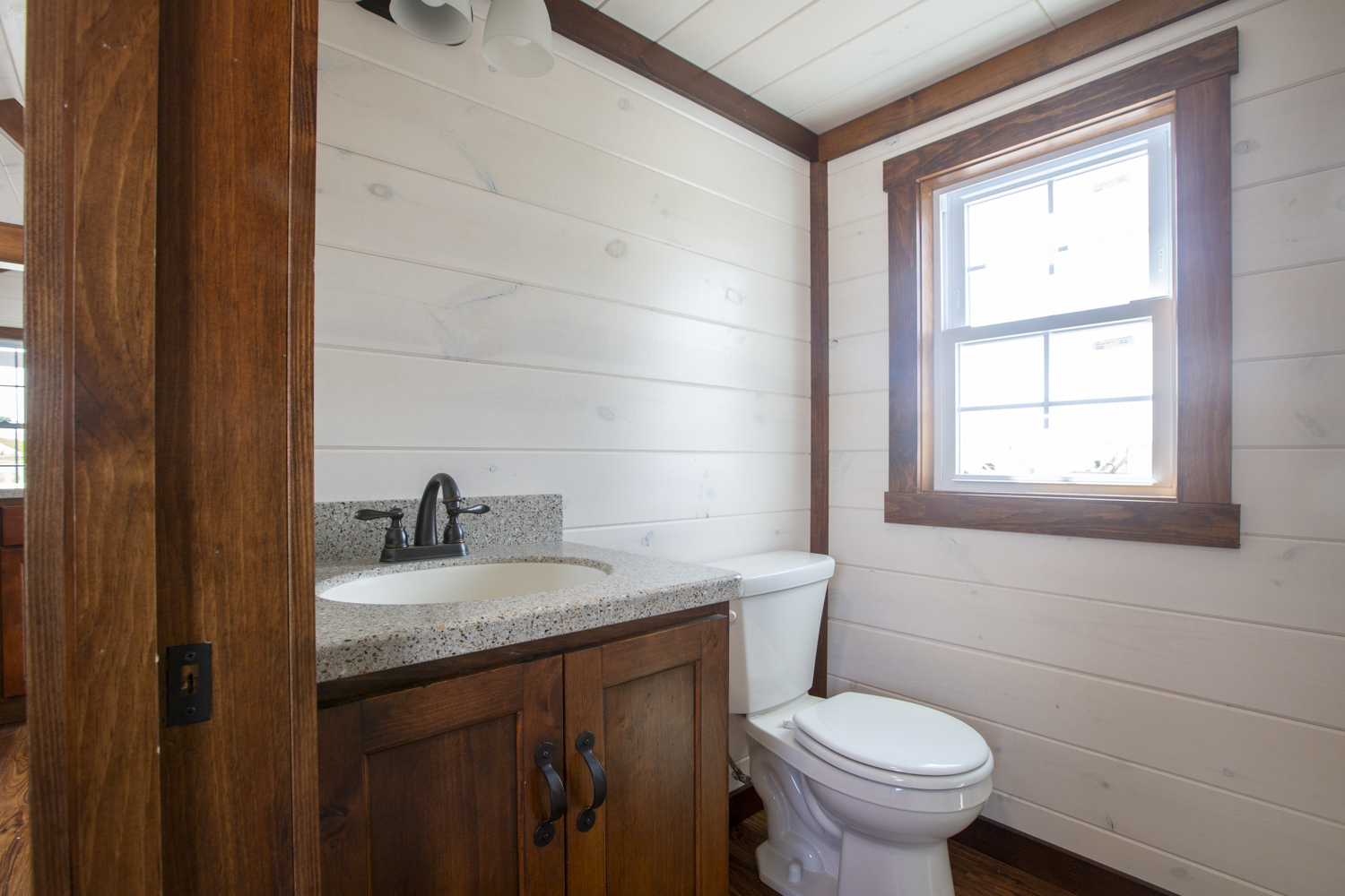 A bathroom in a Santa Rosa Park Model with white plank walls, dark vinyl plank flooring, a window, a white toilet, and a dark wood sink vanity with a dark metallic faucet.