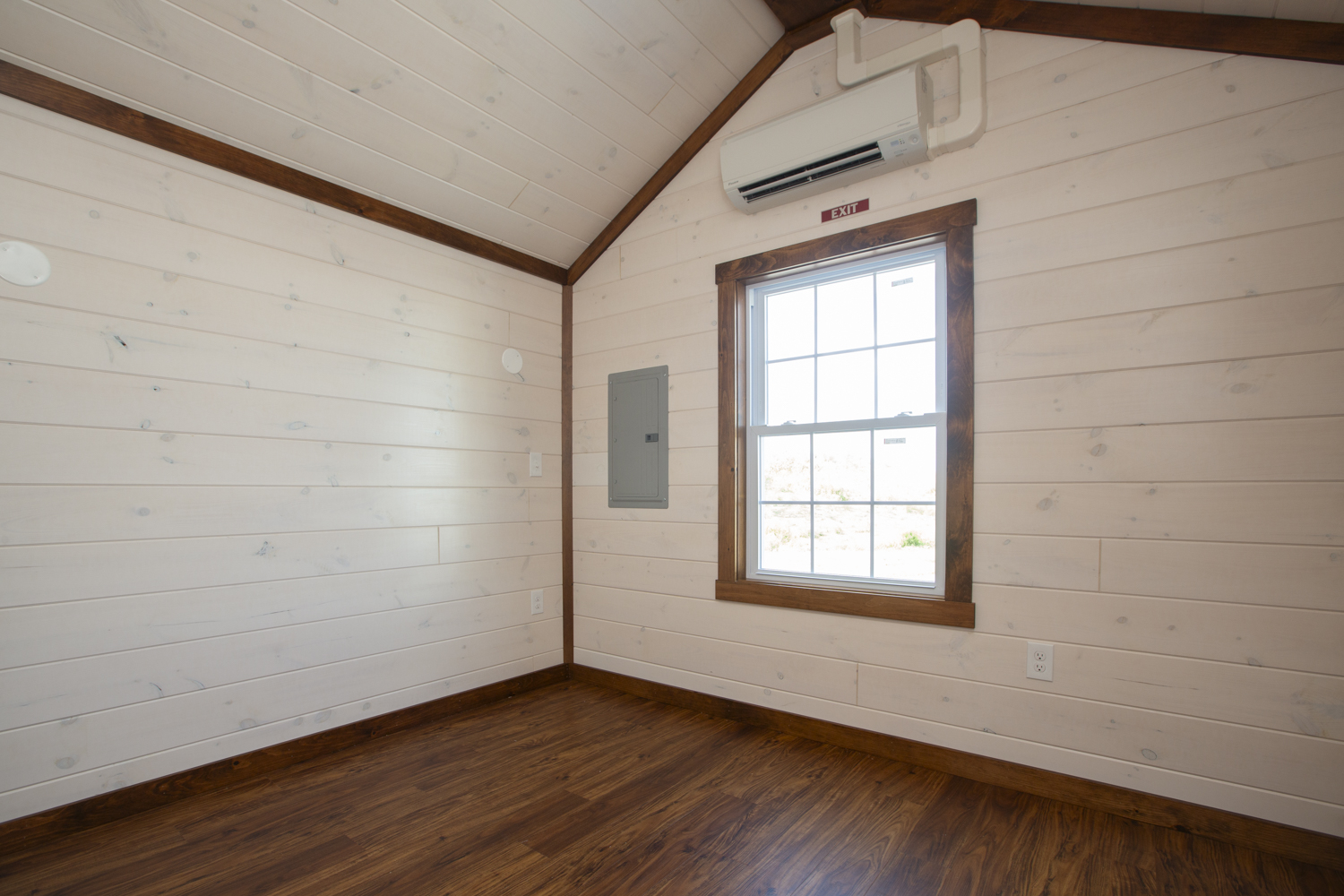 A bedroom in a Santa Rosa Park Model with white plank walls, dark vinyl plank flooring, a large white window with stained wood trim, and a wall-mounted air conditioning unit.