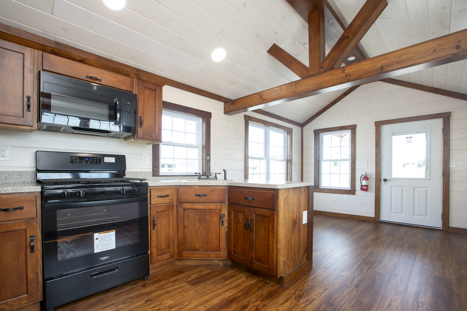 View of a Santa Rosa Park Model kitchen and living room with brown stained wood cabinets, black appliances, a large kitchen sink, white wood plank walls, wood accent beams, and white windows with brown trim.