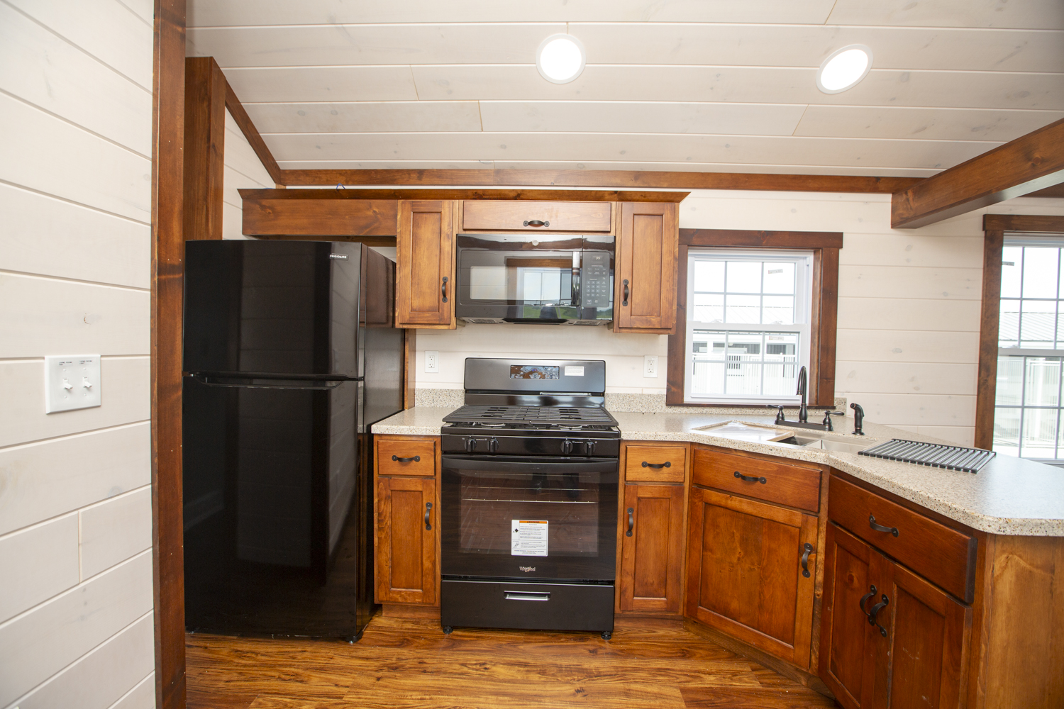 Part of a Santa Rosa Park Model kitchen with brown stained wood cabinets, black appliances, a large kitchen sink, white wood plank walls, and white windows with brown trim.