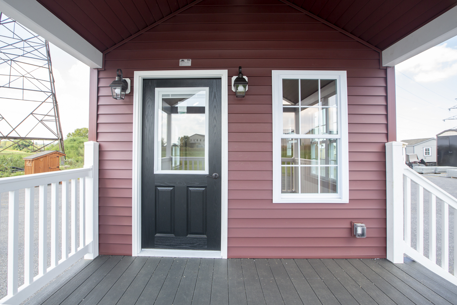 Photo of a Santa Rosa Park Model entrance with red vinyl siding, a white fenced-in front porch, white trim, a white window, and a black front door.