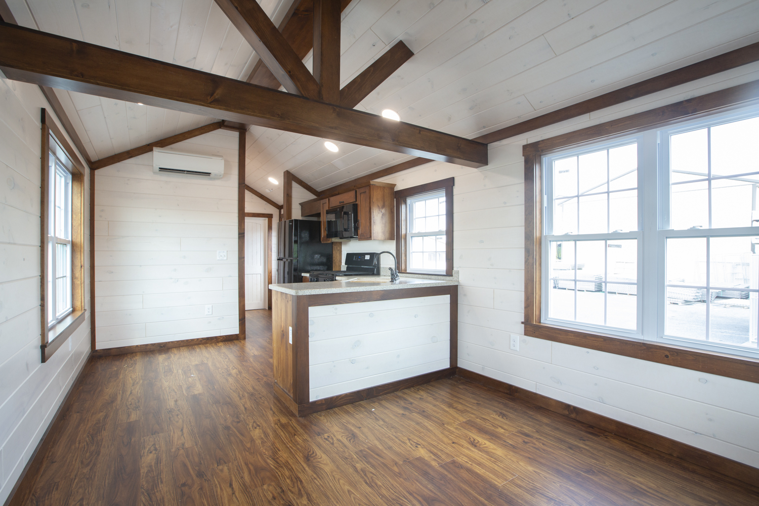 Interior of a Santa Rosa Park Model with wood accent beams, white plank walls, vinyl plank flooring, and a kitchen with black appliances.