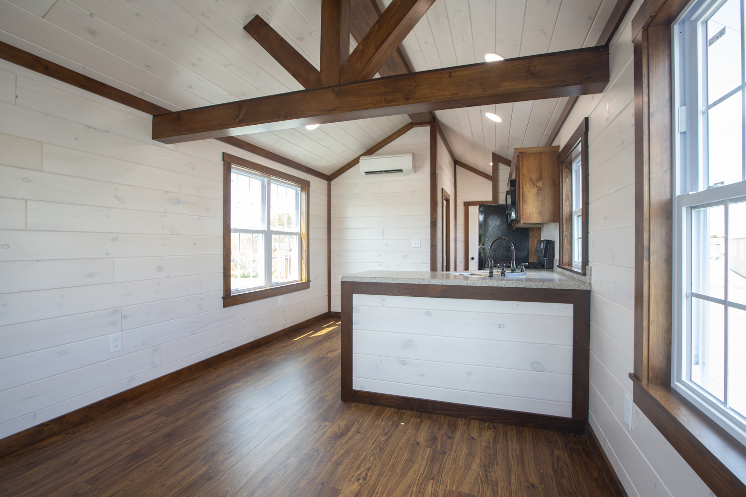 Interior of a Santa Rosa Park Model with wood accent beams, white plank walls, vinyl plank flooring, and a kitchen with black appliances.