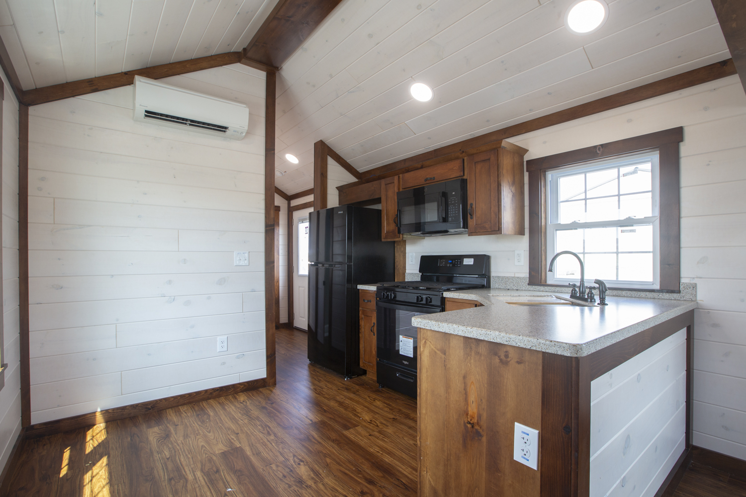 A Santa Rosa Park Model kitchen with dark stained wood cabinets, light colored countertops, black appliances, white plank walls, and dark vinyl plank flooring.