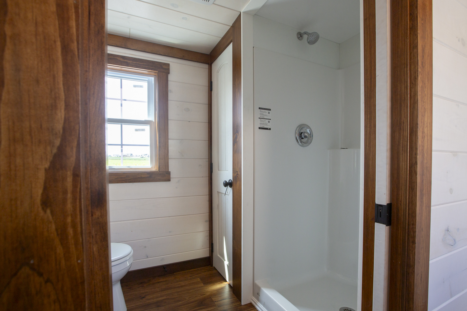 A bathroom in a Santa Rosa Park Model with white plank walls, dark vinyl plank flooring, a white bathroom closet door, a white standing shower, a window, and a white toilet.