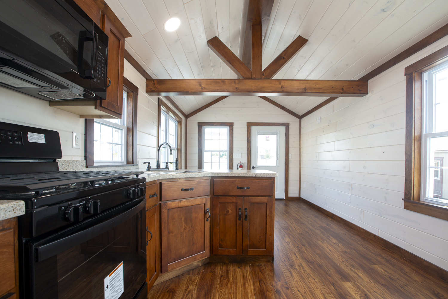 Part of a Santa Rosa Park Model kitchen and living room with brown stained wood cabinets, black appliances, a large kitchen sink, white wood plank walls, wood accent beams, and white windows with brown trim.