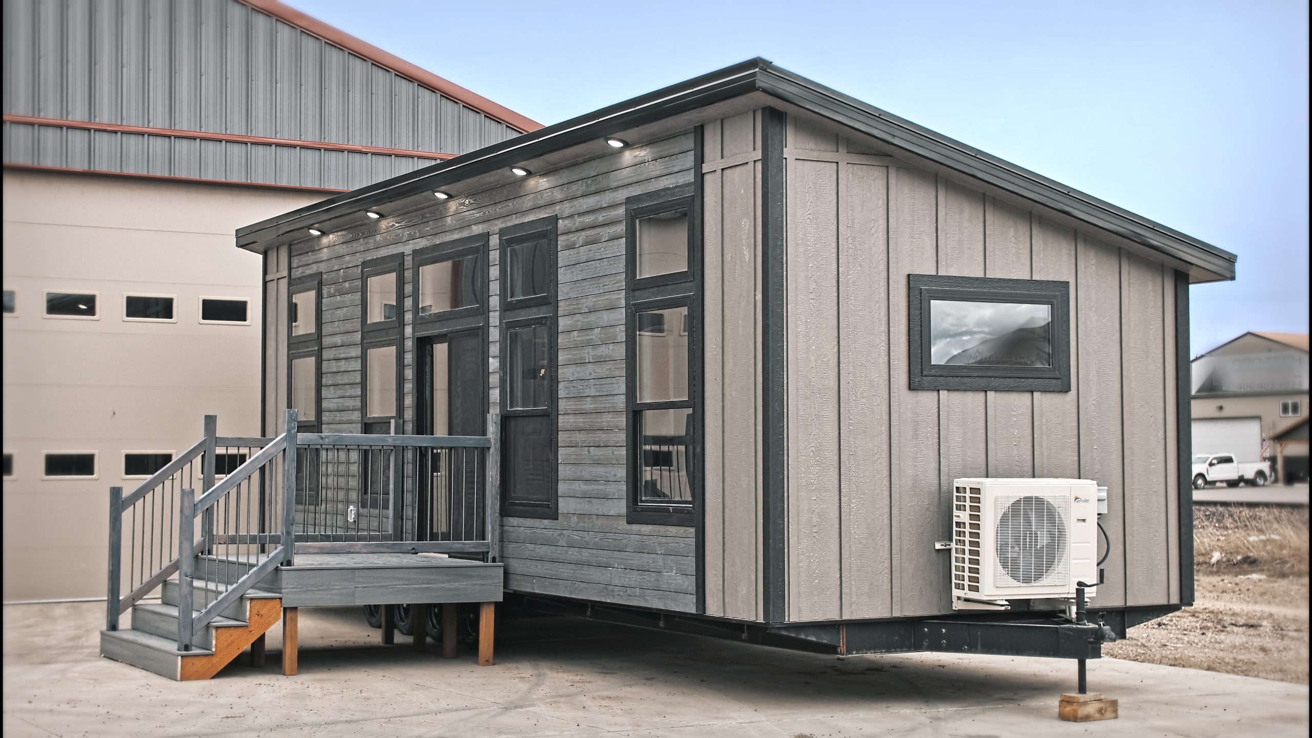 Front and right side exterior of a Meadowlark Park Model on a trailer with gray siding, black trim, a gray wood deck with stairs, and black windows.