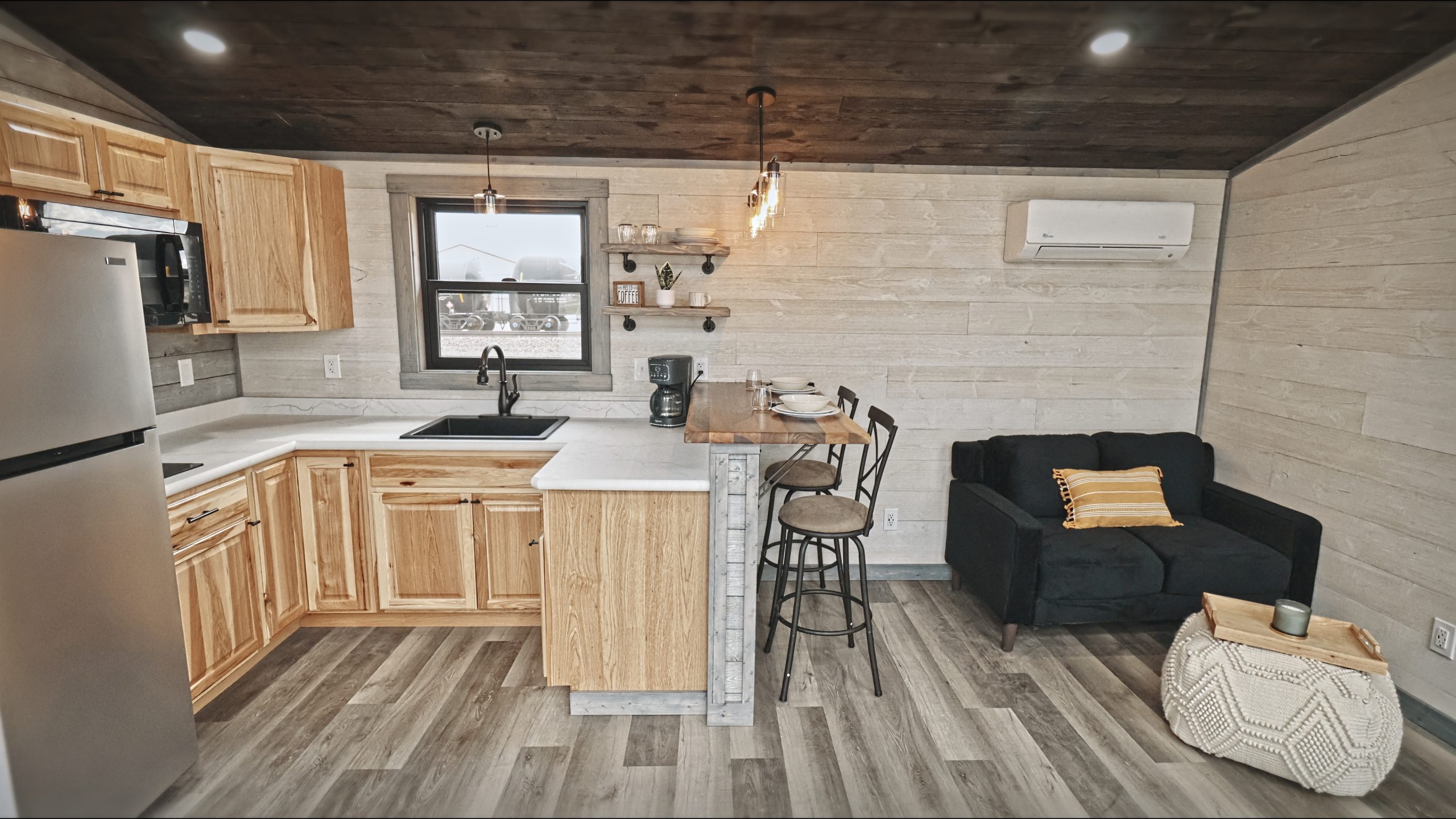 Interior of a Meadowlark Park Model kitchen and living room area with light wood cabinets, white countertops, white plank walls, gray vinyl plank flooring, a brown wood ceiling, and a black loveseat.