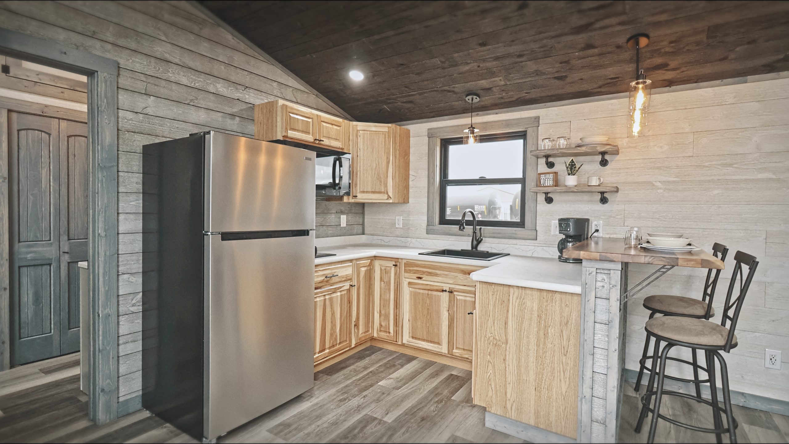 Interior of a Meadowlark Park Model kitchen with light wood cabinets, a stainless steel fridge, white countertops, open shelves, white plank walls, gray vinyl plank flooring, a brown wood ceiling, and 2 bar stools.