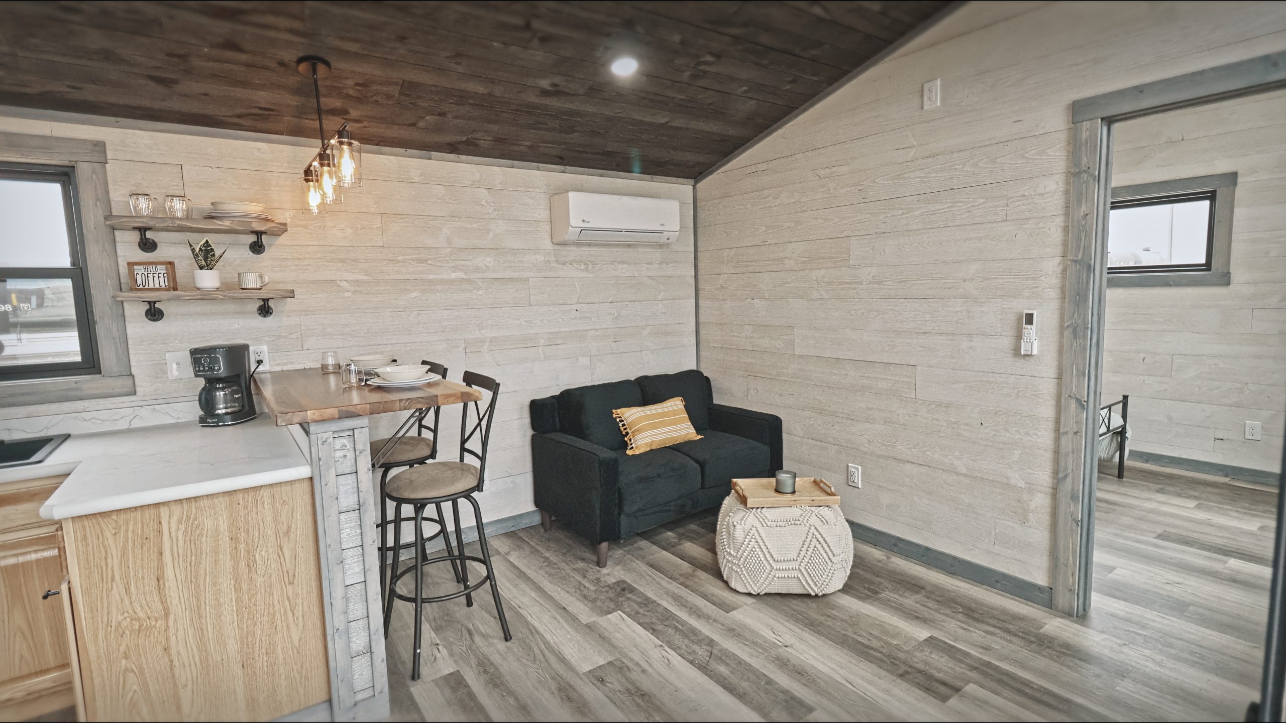 Interior of a Meadowlark Park Model living room area with white plank walls, gray vinyl plank flooring, a brown wood ceiling, a black loveseat, a white pouf cushion, and a dining area with 2 bar stools.