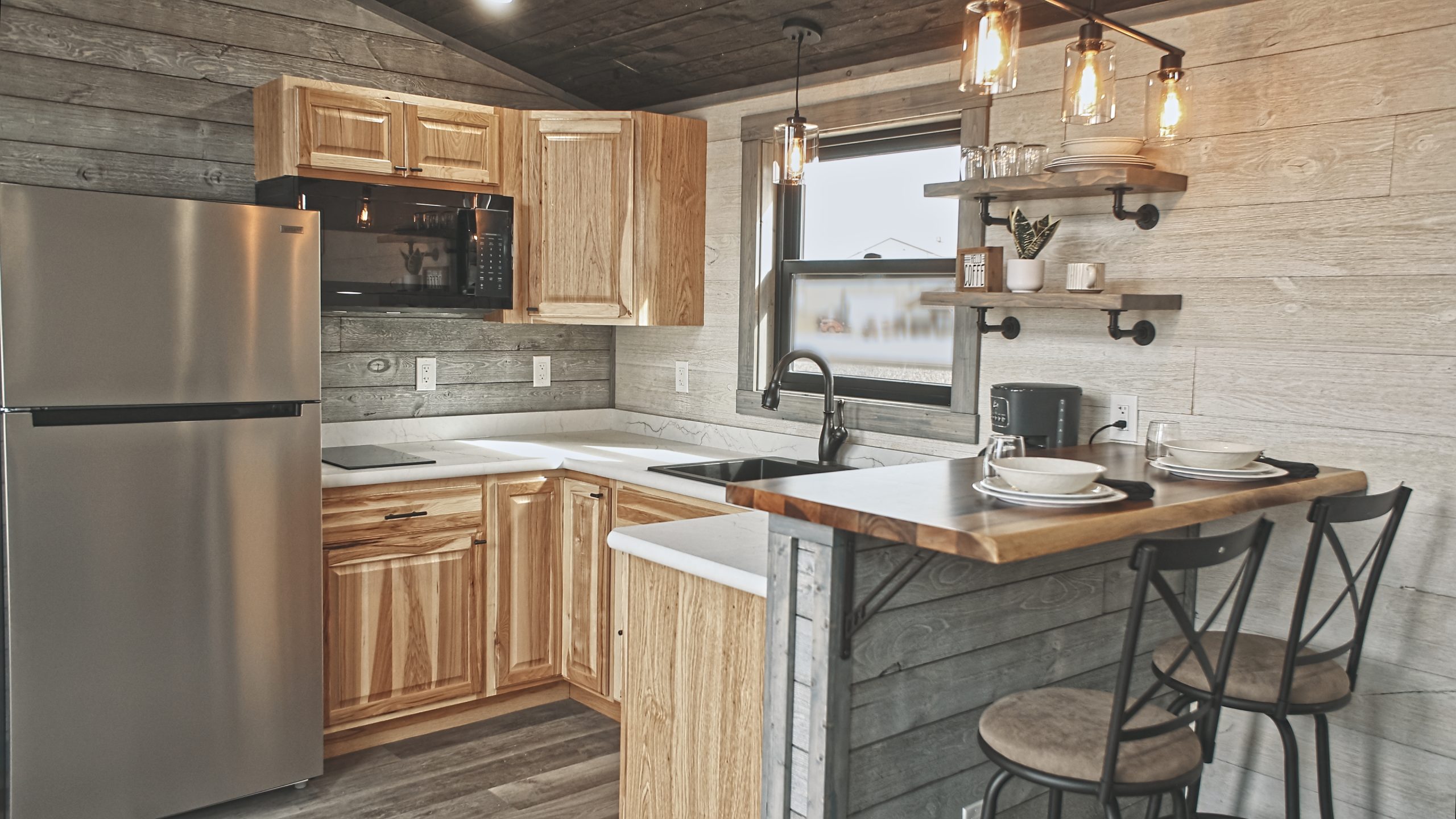 Interior of a Meadowlark Park Model kitchen with light wood cabinets, a stainless steel fridge, a black microwave, white countertops, open shelves, white and gray plank walls, and a dining area with 2 bar stools.