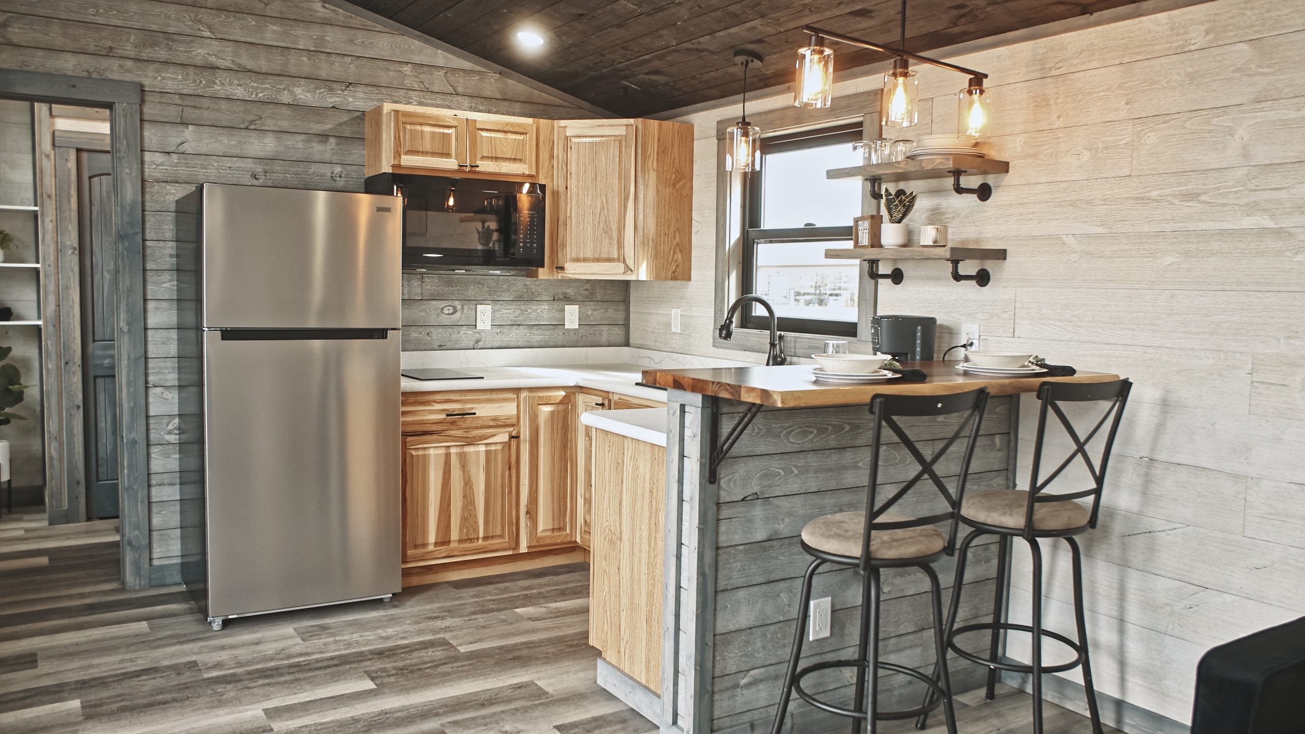 Interior of a Meadowlark Park Model kitchen with light wood cabinets, a stainless steel fridge, a black microwave, white countertops, open shelves, white and gray plank walls, gray vinyl plank flooring, and a dining area with 2 bar stools.