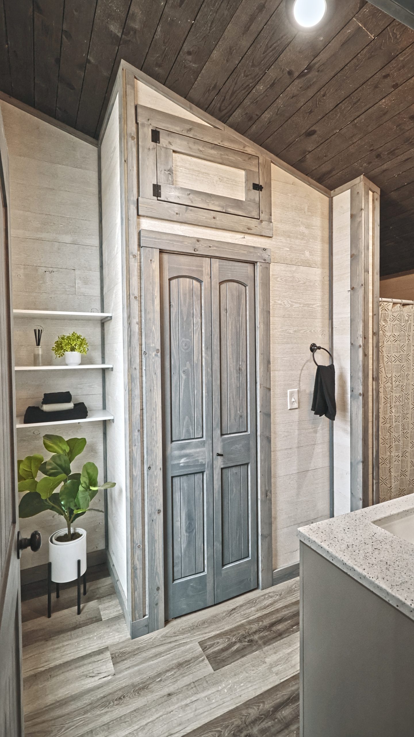 A bathroom in a Meadowlark Park Model with light wood plank walls, gray vinyl plank flooring, white open shelves, a plant, and a gray wood bathroom closet.