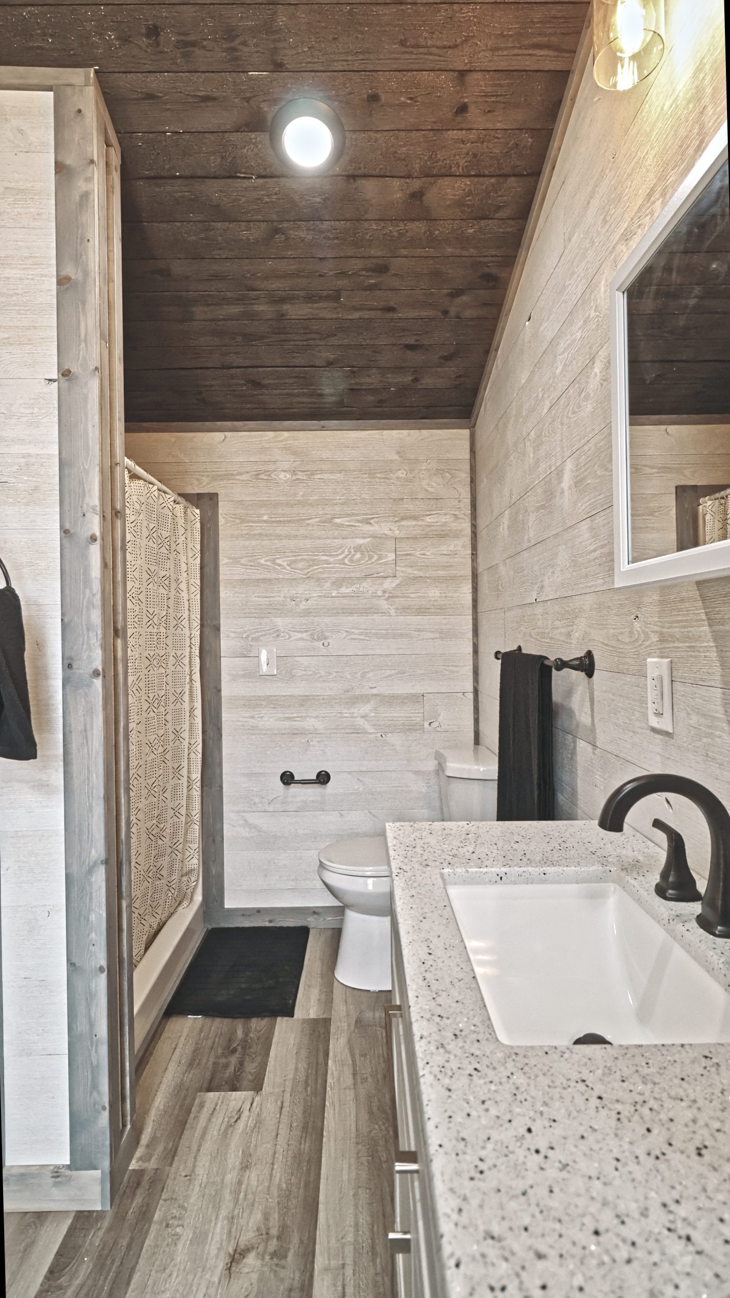 A bathroom in a Meadowlark Park Model with light wood plank walls, gray vinyl plank flooring, a brown wood ceiling, a sink vanity with a mirror, a white toilet, and a shower with a curtain.