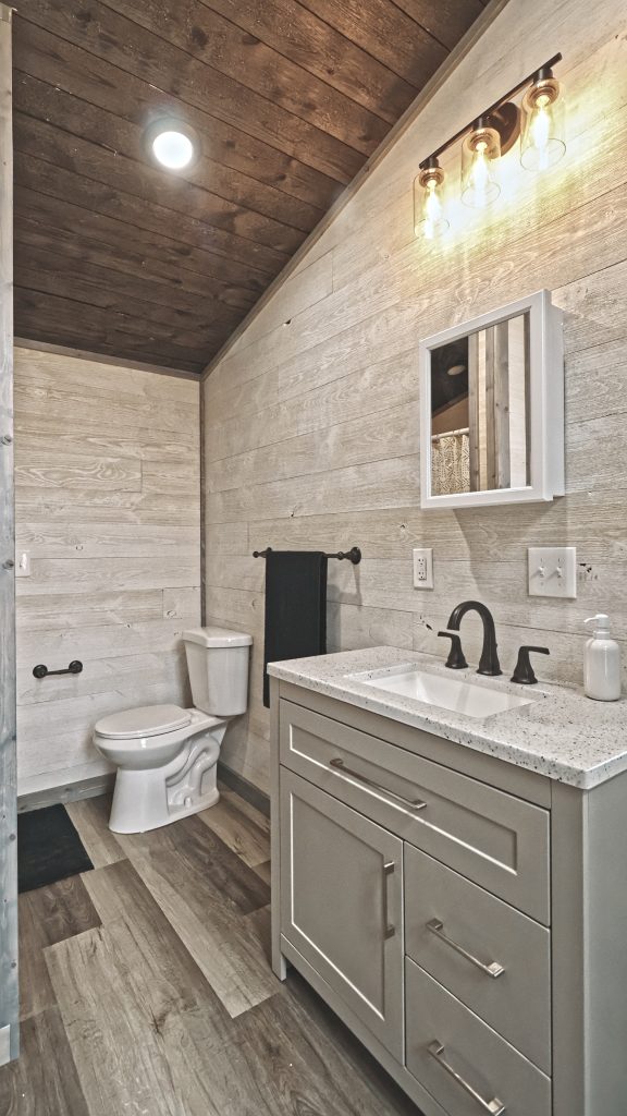 A bathroom in a Meadowlark Park Model with light wood plank walls, gray vinyl plank flooring, a brown wood ceiling, a sink vanity with a mirror, a white toilet, and vanity lighting.