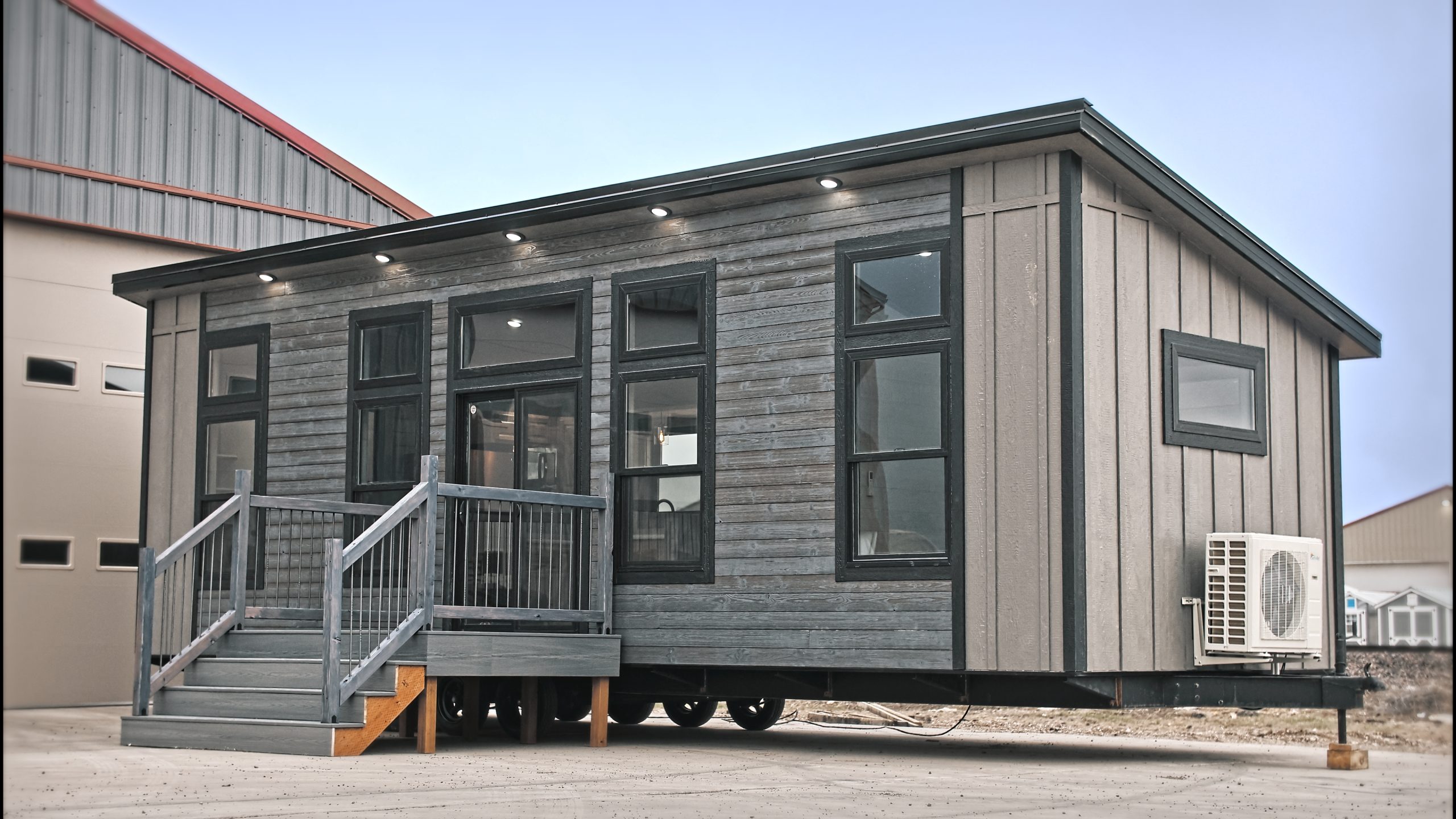 Front and right side exterior of a Meadowlark Park Model on a trailer with gray siding, black trim, a gray wood deck with stairs, and black windows.