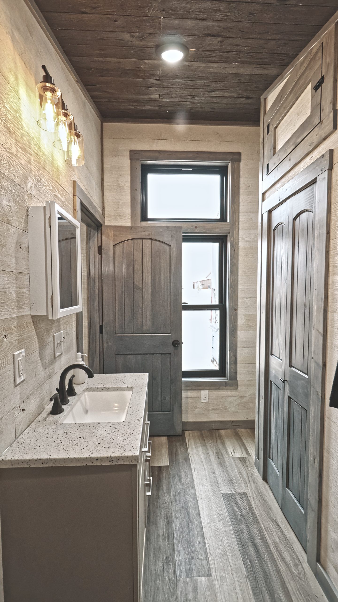 A bathroom in a Meadowlark Park Model with light wood plank walls, gray vinyl plank flooring, a brown wood ceiling, a sink vanity with a mirror, a large black window, and a gray wood bathroom closet.