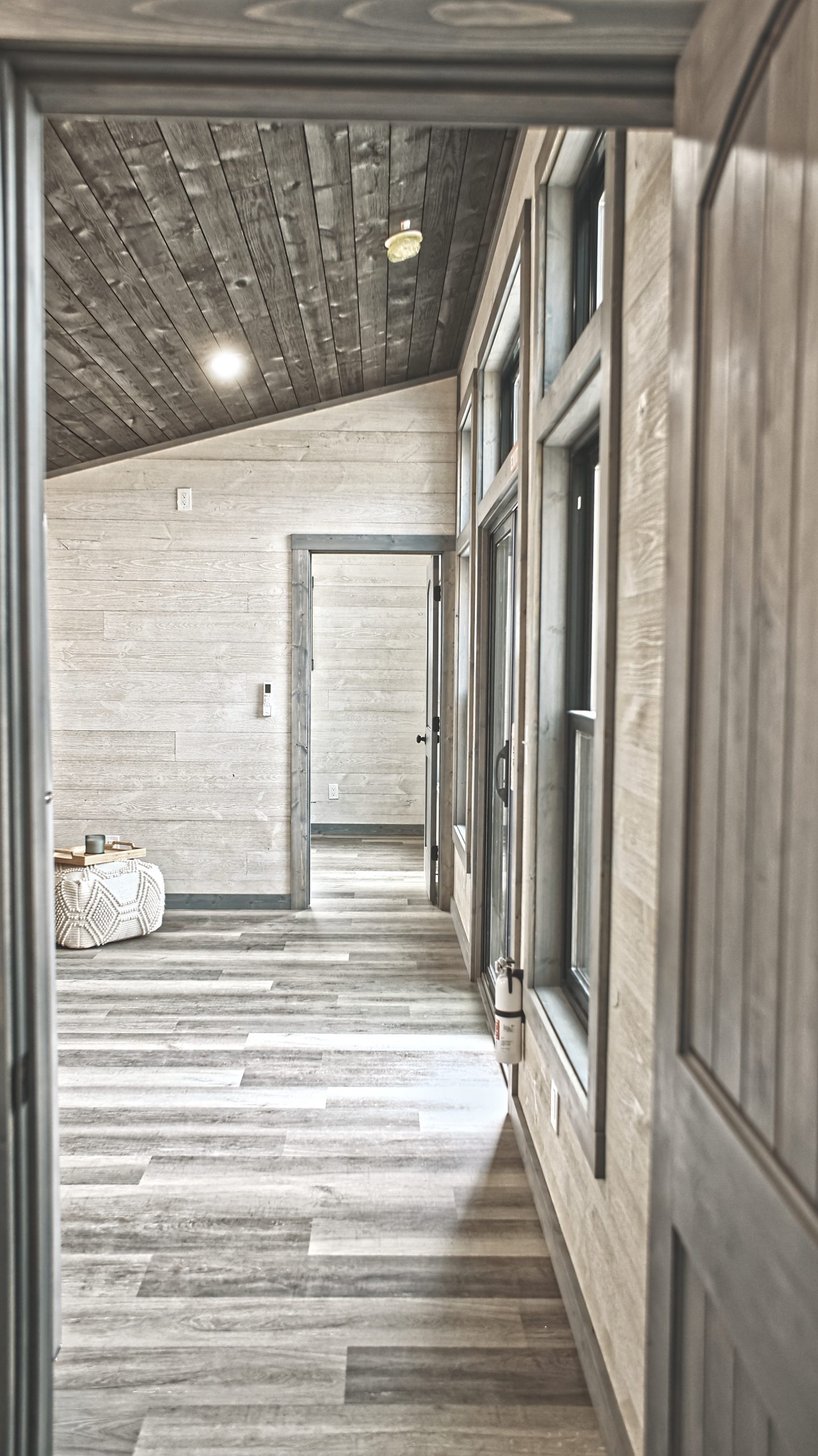 Hallway in a Meadowlark Park Model leading into a room with light plank walls, gray vinyl plank flooring, gray trim, a gray door, and a wood ceiling.