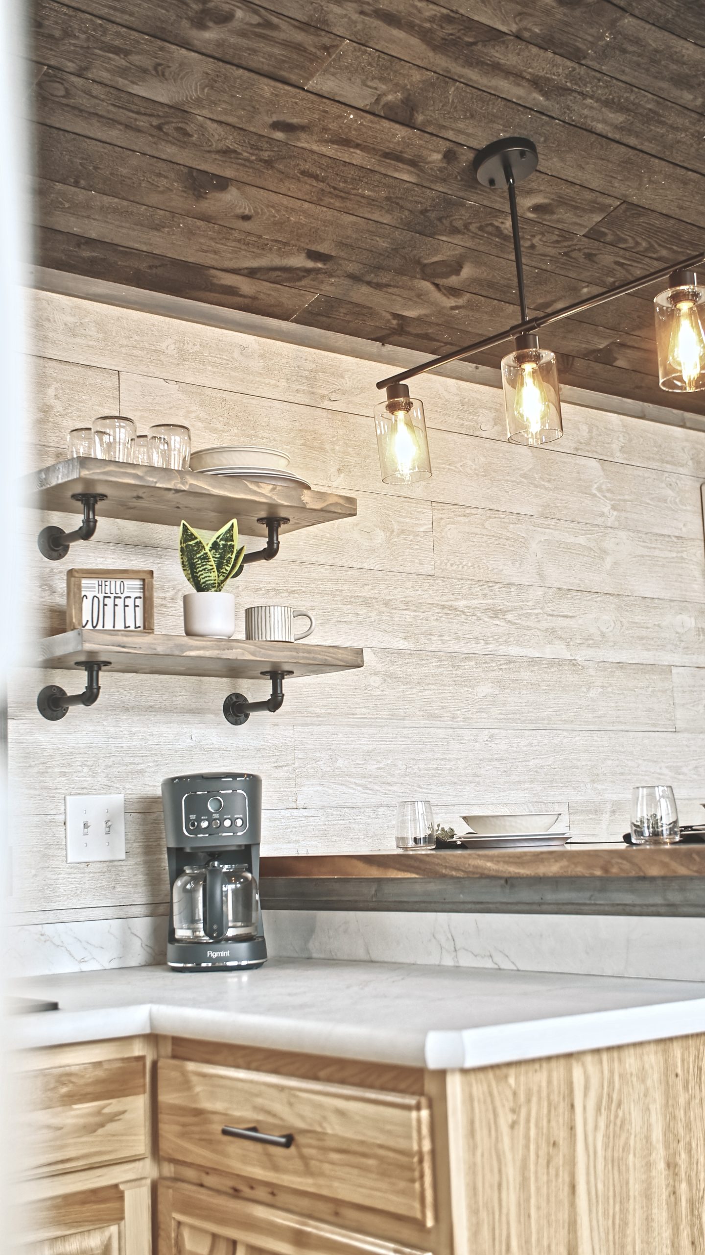 Close up of details in a Meadowlark Park Model kitchen with open shelves, 3-light pendant lighting, light wood cabinets, a coffee maker, and white countertops.