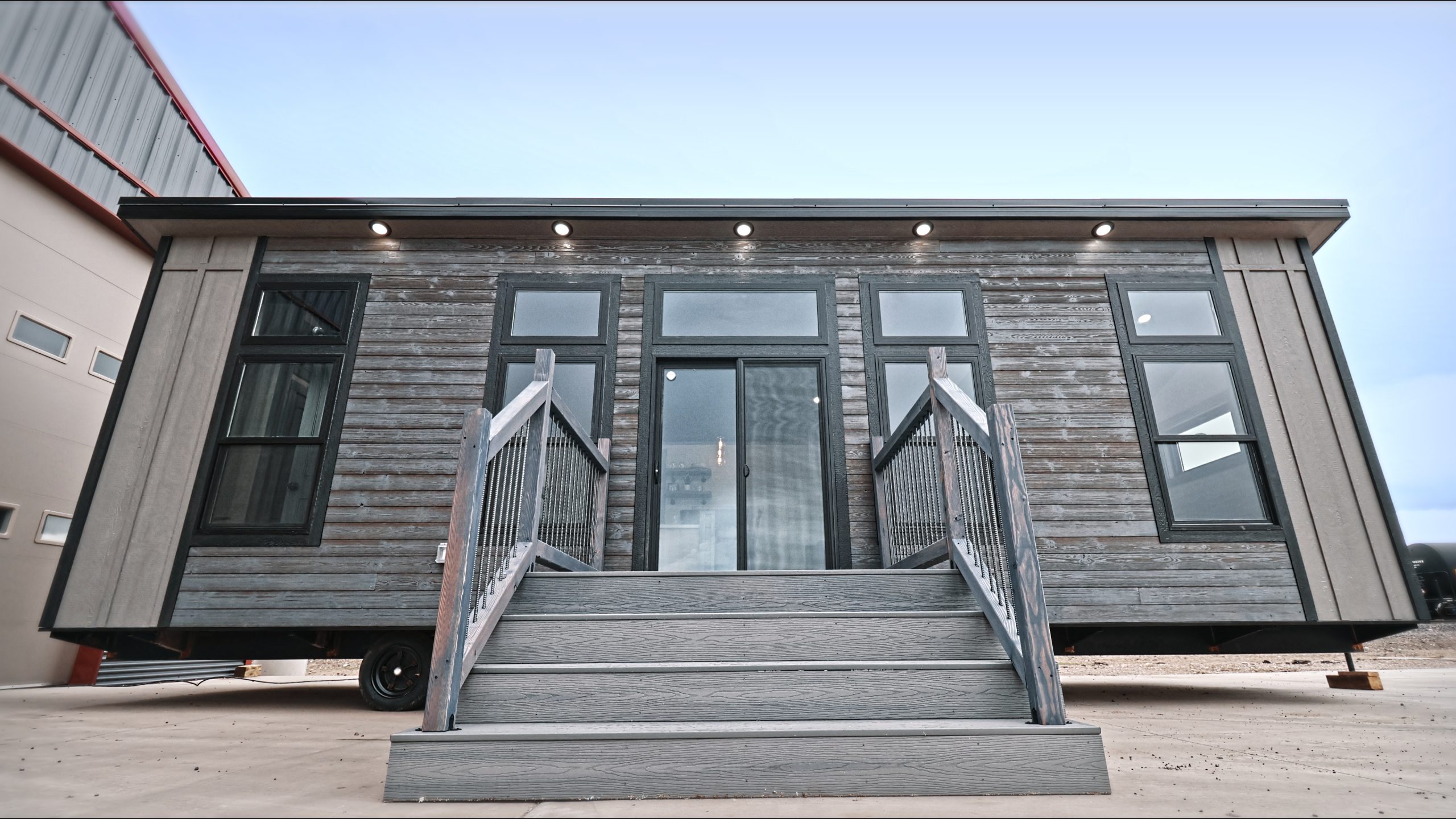 Front exterior and entrance of a Meadowlark Park Model on a trailer with gray siding, black trim, a gray wood deck with stairs, and black windows.