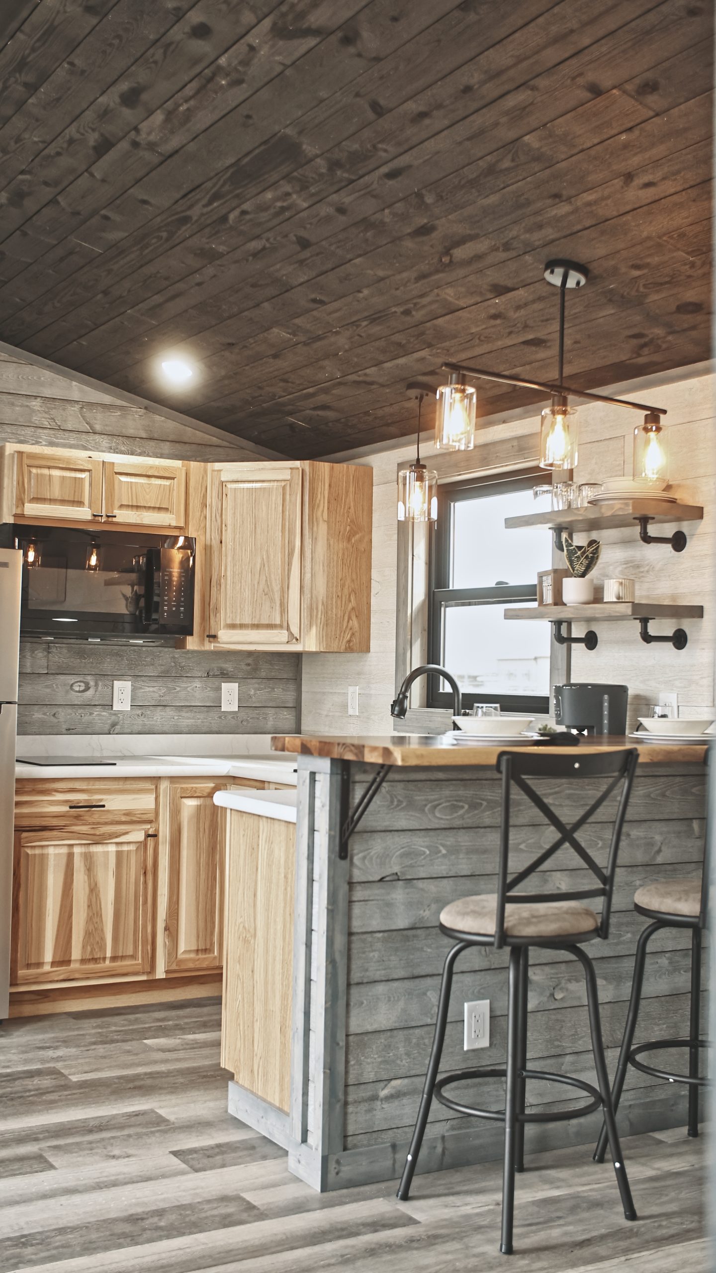 Interior of a Meadowlark Park Model kitchen with light wood cabinets, a black microwave, white countertops, open shelves, white and gray plank walls, and a dining area with 2 bar stools.