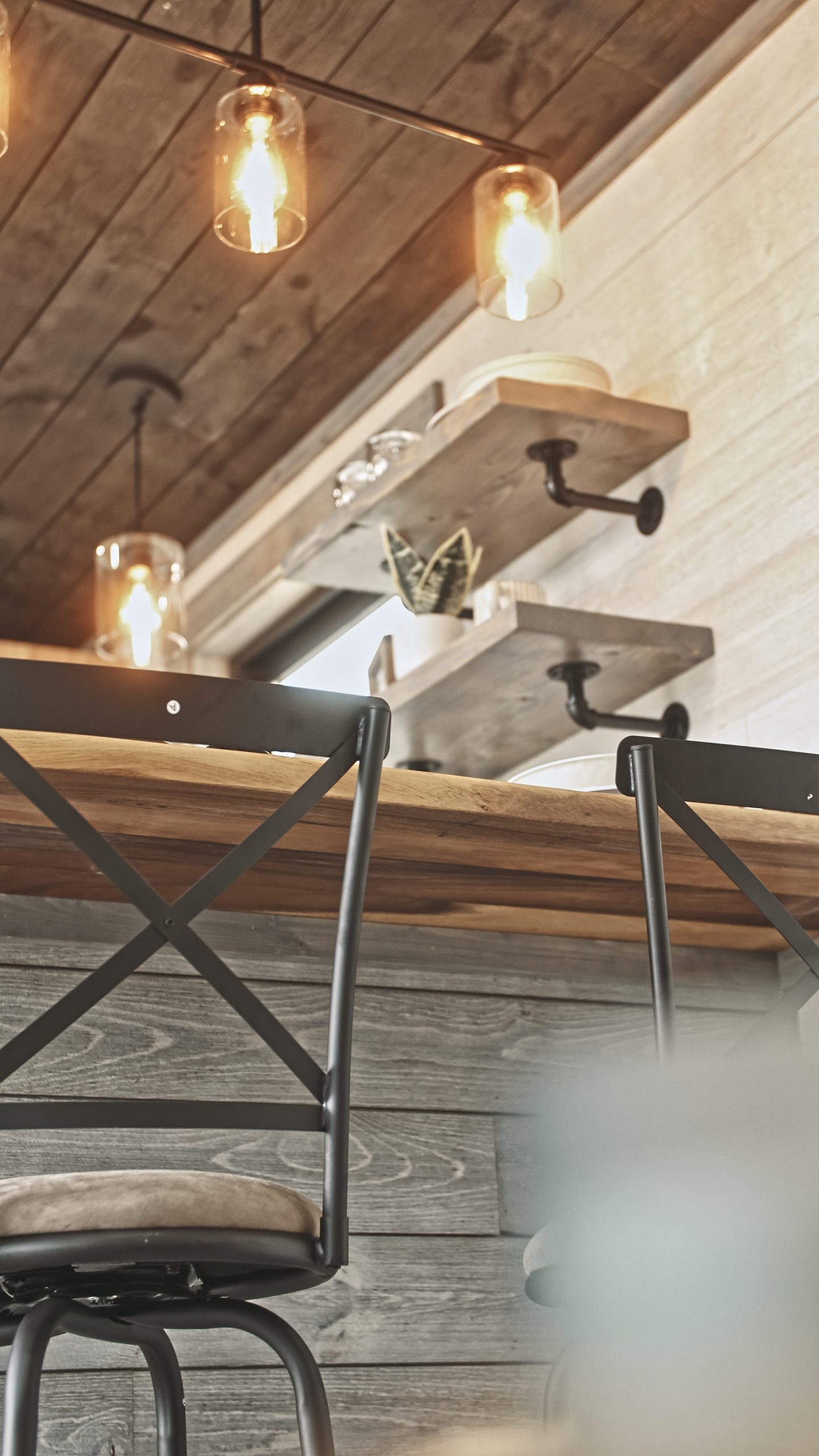 Close up of details in a Meadowlark Park Model kitchen with open shelves, pendant lighting, a dining counter with 2 bar stools, gray and white plank walls, and a brown wood ceiling.