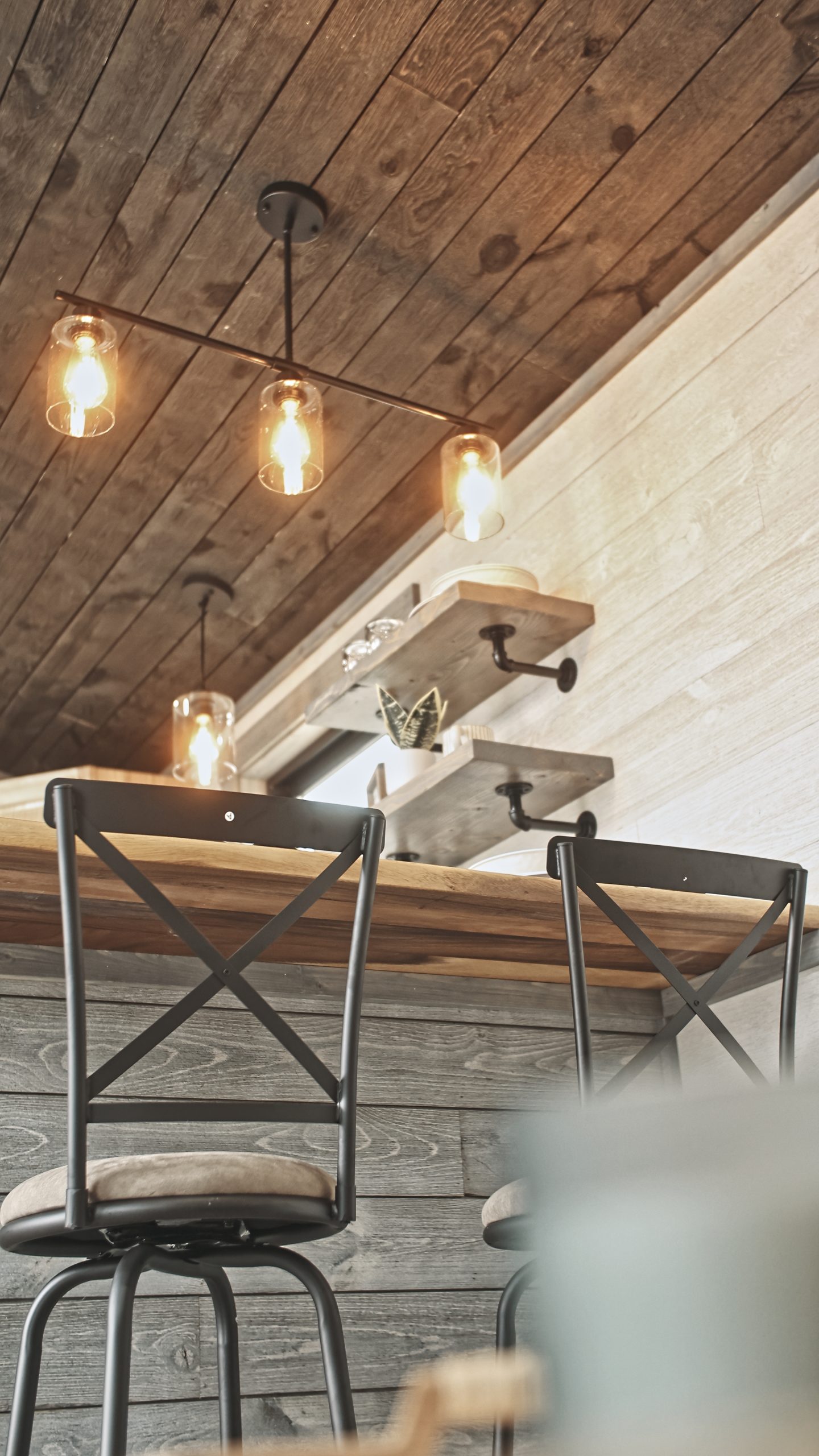 Close up of details in a Meadowlark Park Model kitchen with open shelves, pendant lighting, a dining counter with 2 bar stools, gray and white plank walls, and a brown wood ceiling.